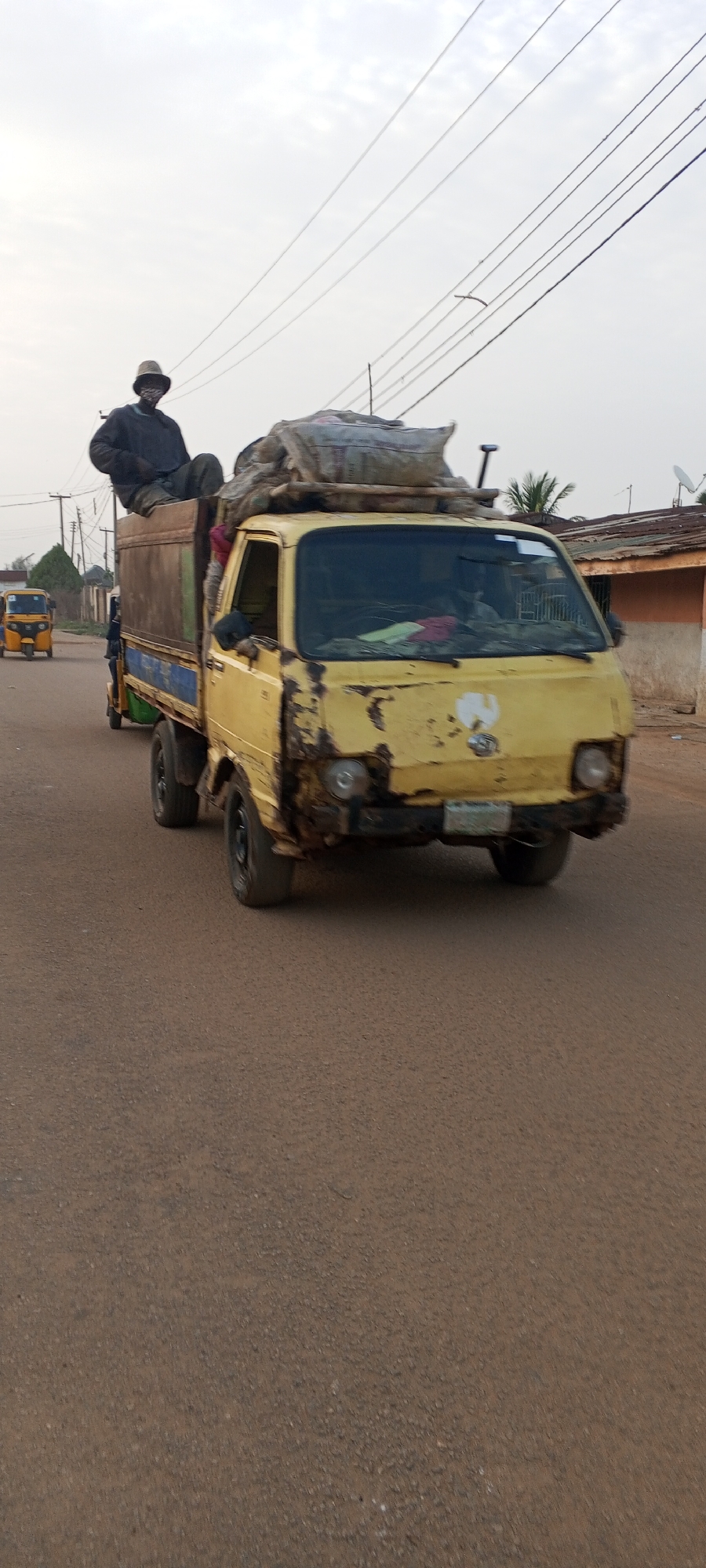 The image shows a yellow, heavily worn pickup truck traveling down a paved road in Sabon Tasha, Nigeria. The truck bed is loaded with items covered by a tarp or similar material, and a man is sitting on top of the load. A second person is in the vehicle. A tricycle taxi is in the background on the left side of the road. Utility poles and lines are visible overhead. Buildings and vegetation line the side of the road. The image is captured during daylight hours.
