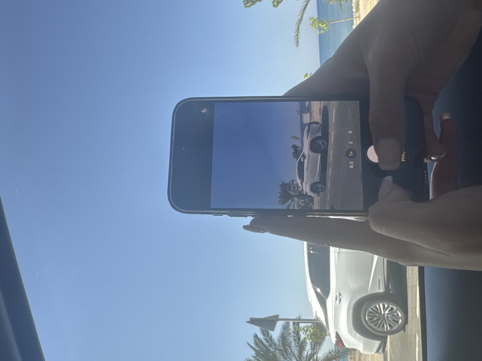 A person is holding a smartphone, capturing an image of a white Lexus RX 350 parked near a body of water. The car is a modern luxury SUV with a sleek design, featuring chrome alloy wheels and a distinctive grille. In the background, palm trees and lush greenery suggest a warm, tropical climate. The sea stretches out towards the horizon under a clear blue sky. The scene appears to be a coastal road in Polis Chrysochous, Cyprus, known for its natural beauty and tranquil atmosphere. The car is positioned on the right side of the frame, while the sea and sky dominate the left. The image is taken from inside a vehicle, likely a car, as indicated by the dashboard and window frame.