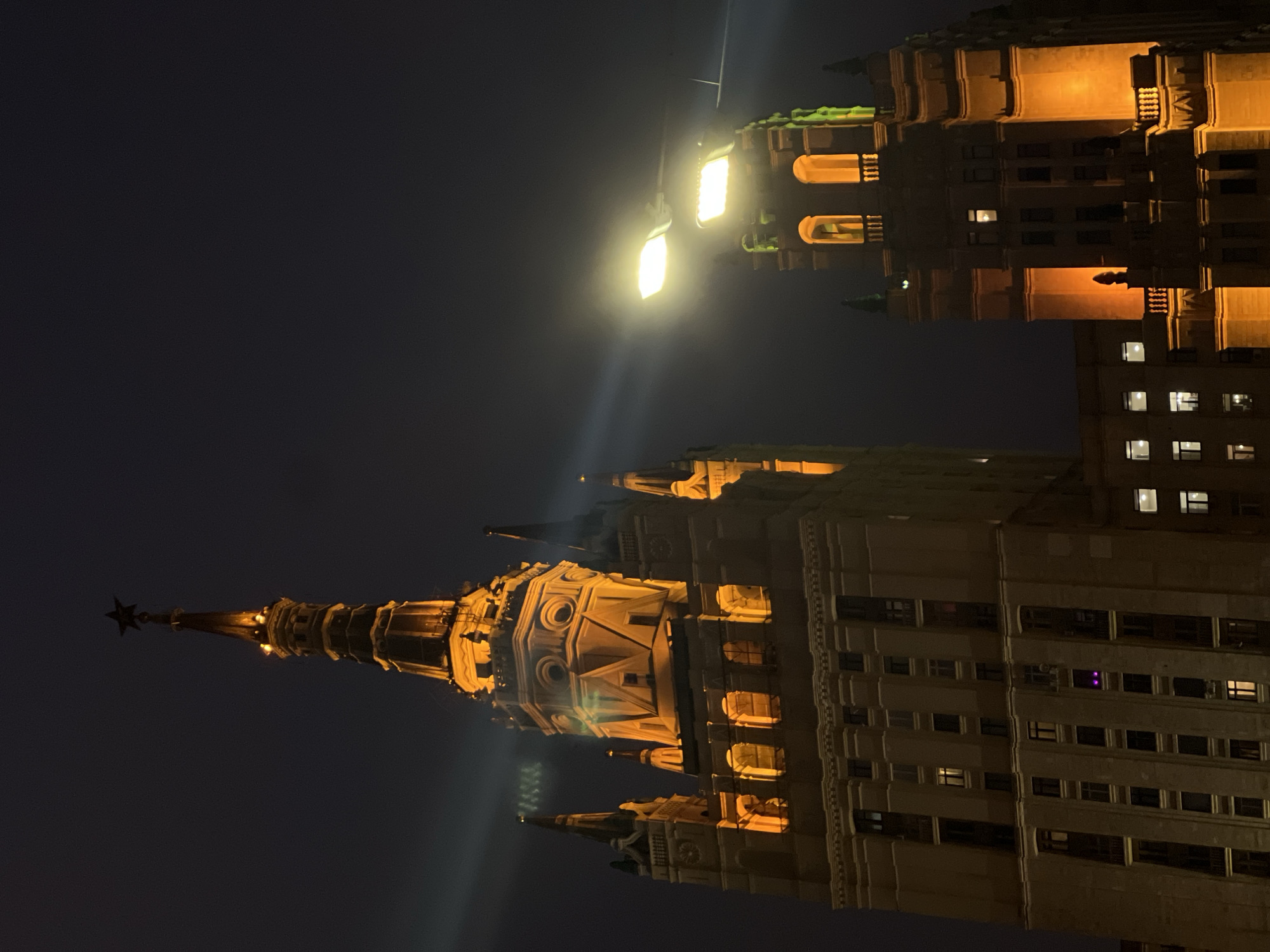 At night in Moscow, Russia, illuminated Stalinist architecture is seen under a dark sky. The primary building, a skyscraper featuring a spire topped with a red star, is lit with warm yellow light. The building exhibits multiple levels and ornate details, including recessed windows and decorative architectural elements. A street light shines brightly in the upper portion of the frame, causing lens flares. A second building, similarly lit and styled, is seen in the background, with lit windows visible.
