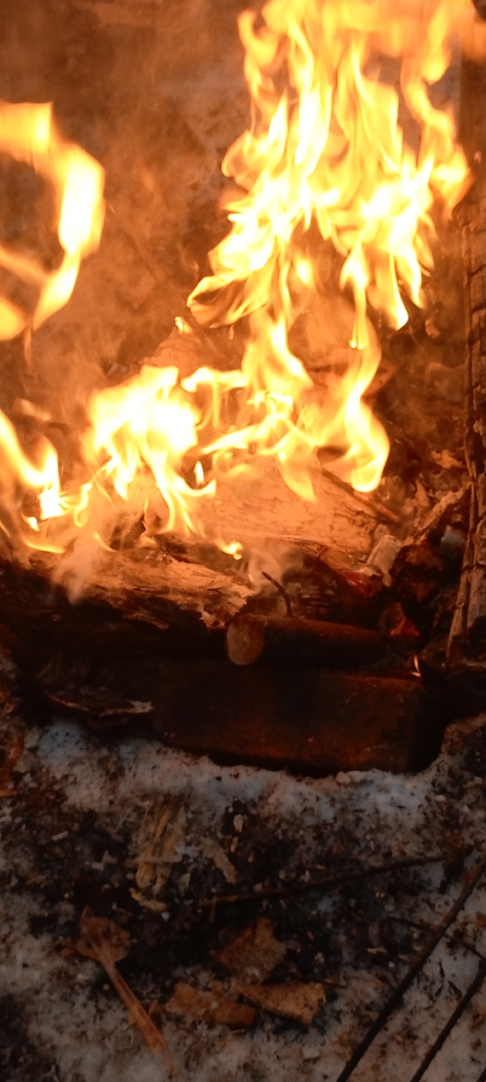 A bonfire roars with bright yellow flames. Logs are piled within the fiery heart, surrounded by wisps of smoke. Below, ash and remnants of previous burns mix with patches of snow. The ground is a blend of dark char and cold white. The composition captures the contrast of fire and ice.