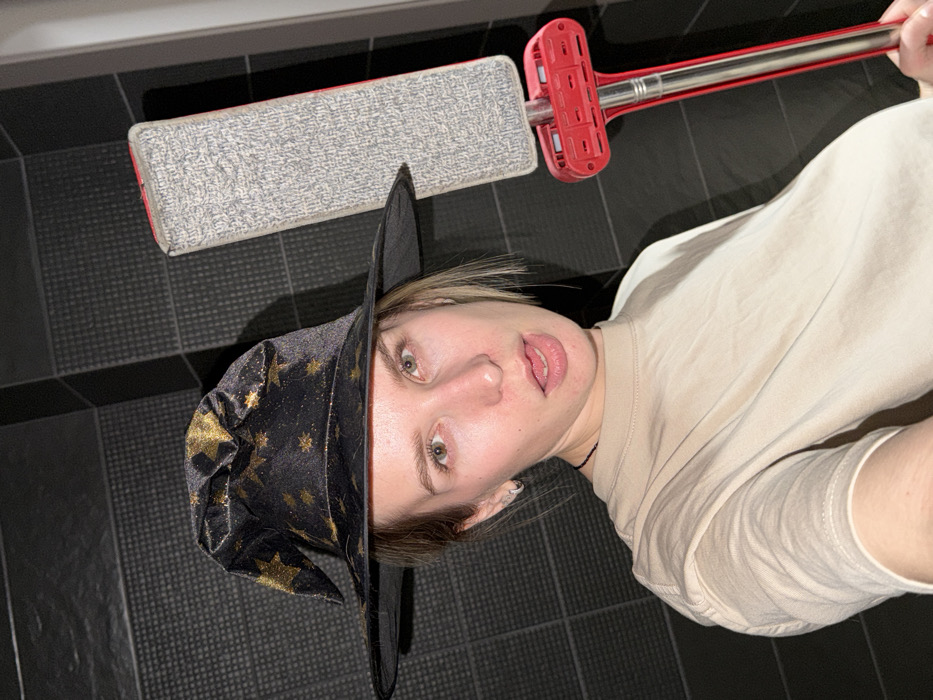 An indoor portrait displays a young adult female subject, positioned centrally and looking directly at the camera. She wears a black pointed witch hat adorned with gold stars and a plain beige short-sleeved t-shirt. Her light-colored hair frames her face, and her eyes are light-colored. A small silver hoop earring is visible in her left ear. Her mouth is slightly open, with her tongue partially extended. To her right, she holds a flat mop vertically, featuring a rectangular grey microfiber head, a red base, and a silver-colored handle. The background consists of dark, possibly black, square or rectangular tiles arranged in a grid pattern. A vertical lighter-colored line, consistent with a doorframe or wall edge, is present on the far right. The image contains no discernible visual identifiers for a specific location within Chaikovsky, Russia.