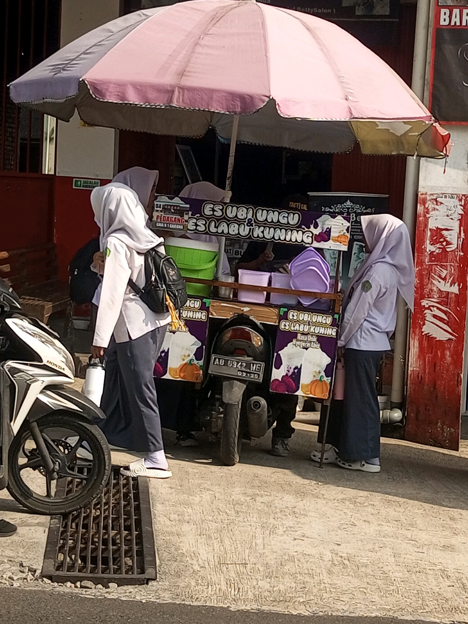 A street vendor in Krasak, Indonesia, is selling ice cream from a cart parked next to a motorcycle. The cart is adorned with purple and white signage displaying the words "ES UBI UNGU ES LABU KUNING" and images of what appear to be purple sweet potato and pumpkin. Two young women wearing headscarves and school uniforms are standing near the cart. One is reaching for something on the cart, while the other stands behind her. The scene takes place on a sunny day, with the vendor's cart and the surrounding pavement illuminated by sunlight. The background shows a building with red and white accents, and a portion of a motorcycle's front wheel is visible in the foreground.