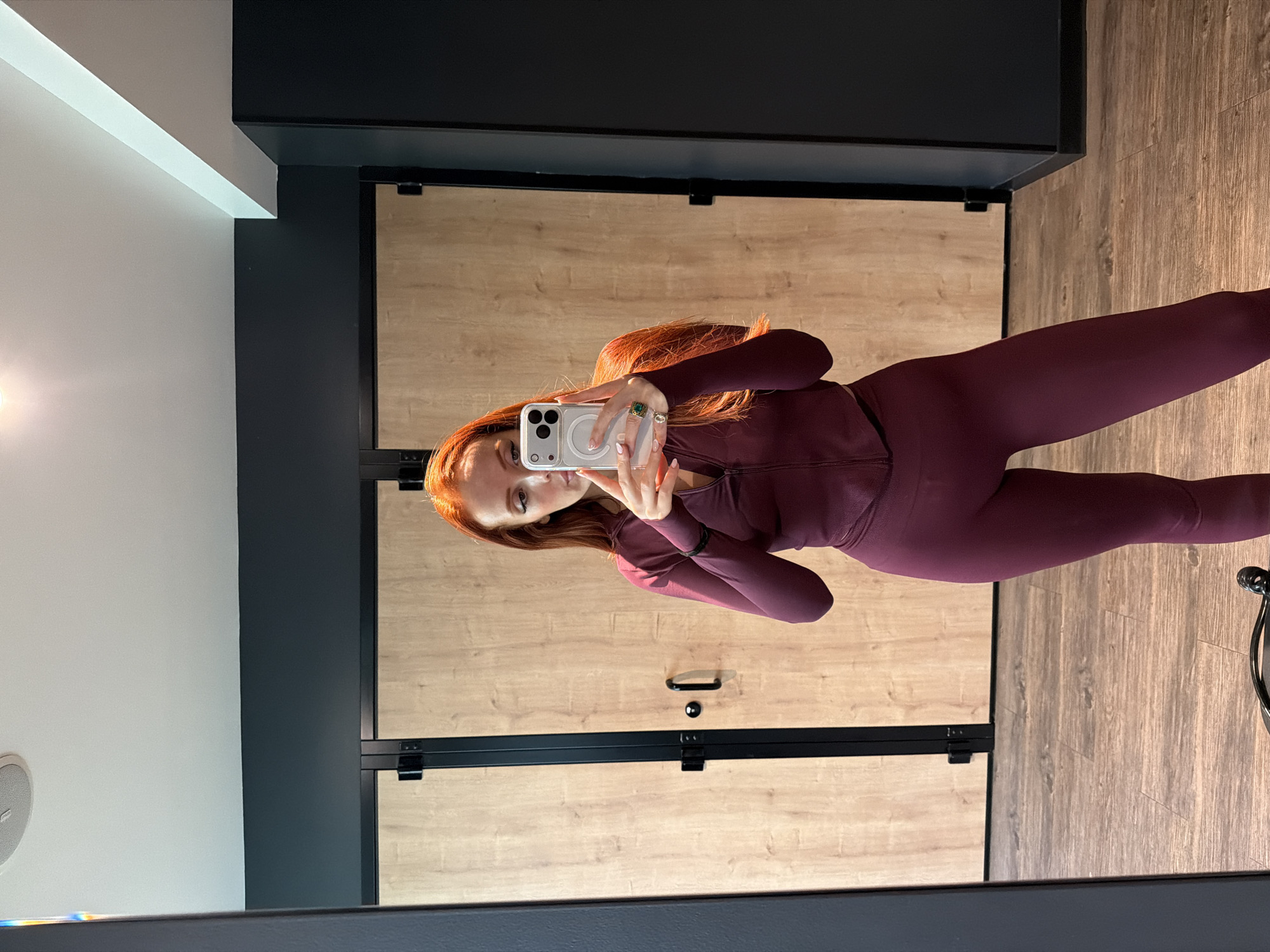 A woman with long, red hair is taking a selfie in front of a mirrored wall. She is wearing a burgundy athletic set. The wall behind her consists of a light wood pattern, divided into panels by black metal framing. A black frame surrounds the mirror in which the woman is visible. Above the mirror is a dark gray surface, likely a shelf or a part of the building's architecture. A white ceiling is partially visible on the left side of the frame, next to a gray wall. The location cannot be determined based on the image.