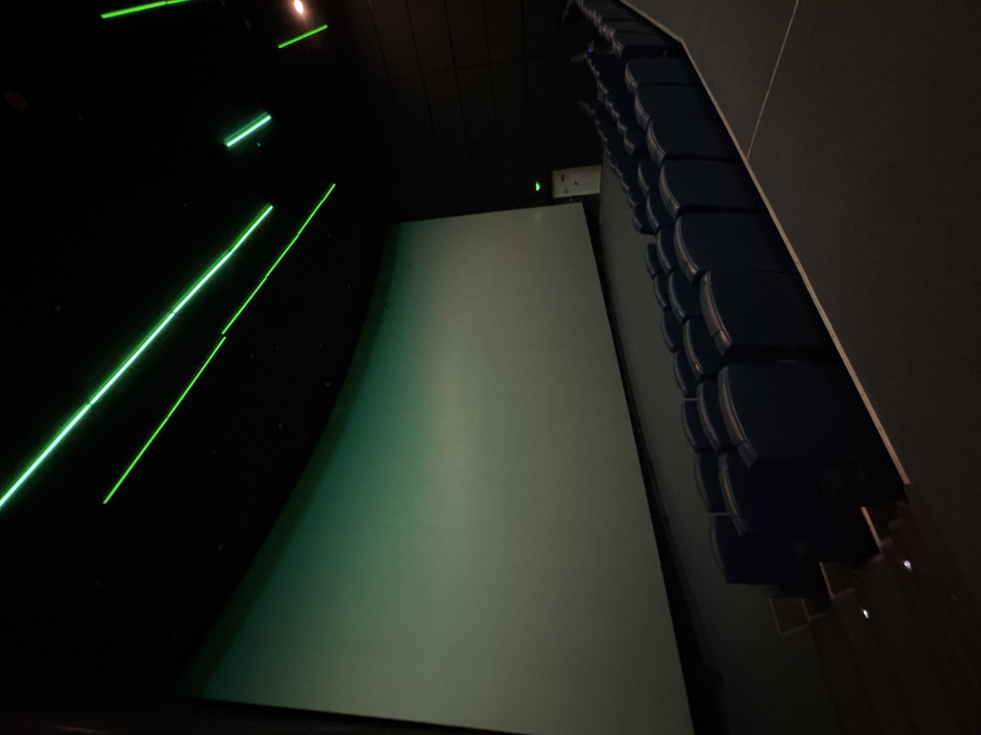 The image depicts the interior of a movie theater. A large screen dominates the scene, with a noticeable green hue across its surface. To the right, rows of dark blue seats are visible. The left side of the image is predominantly black, with thin, bright green lines running diagonally across it. The overall lighting is dim, contributing to the dark and enclosed atmosphere of the theater.