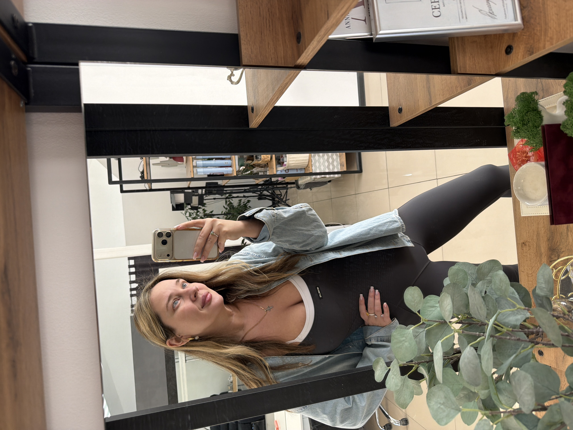 The image captures a mirror selfie of a young woman taken indoors. The woman is positioned upright, facing slightly right, holding a gold-cased iPhone with her right hand to take the picture, reflecting her image in a large mirror. She has long, straight blonde hair and blue eyes, looking towards the camera with a subtle smile. She wears a light blue denim jacket over a dark gray or black form-fitting bodysuit with a visible white sports bra or lining underneath. A delicate gold necklace with a small cross pendant is visible around her neck. Multiple rings adorn her fingers, and her nails are manicured with a natural, light polish.
The setting appears to be a modern interior space, likely in Chaikovsky, Russia, possibly a salon, studio, or professional office, featuring a minimalist design. Behind her, visible in the mirror's reflection, are wooden shelves supported by black metal frames. These shelves display various decorative items, including rolled textiles or paper, baskets, and small ornamental plants. Prominently featured on one shelf is a silver-framed document with the word "CERTIFICATE" and partially visible "ANN..." in English, alongside some Cyrillic script. Other decorative elements include artificial plants, such as eucalyptus-like branches in the foreground, and a vibrant green moss-like object on a shelf. The floor is tiled with light-colored square tiles. The lighting is even, suggesting an indoor artificial light source or bright diffused natural light. The overall impression is one of casual elegance in a contemporary setting.