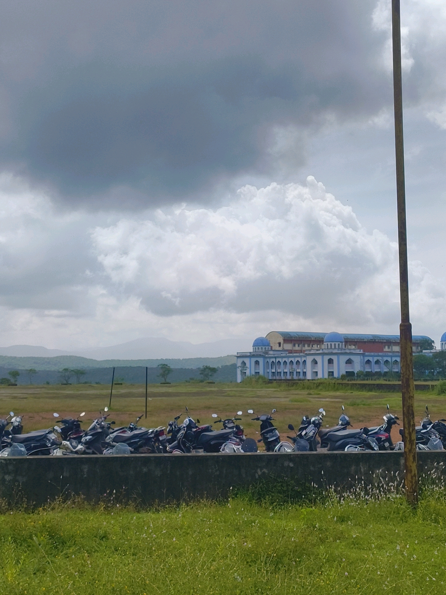 The image captures an outdoor scene in Marukeri, India, under a cloudy sky with large, puffy white clouds dominating the upper portion. In the middle ground, a row of parked motorcycles, predominantly black and silver, is lined up behind a concrete barrier. Beyond the motorcycles, a vast, open field stretches towards a long, white building with blue domes, indicative of a mosque or a similar institution. The building features arched windows and a symmetrical architectural design. In the far background, a range of distant, green hills fades into the hazy sky. To the right of the frame, a vertical, weathered wooden pole stands prominently. The foreground consists of lush green grass with scattered small white wildflowers. The overall atmosphere is overcast and somewhat subdued, possibly suggesting an approaching or departing rain shower.