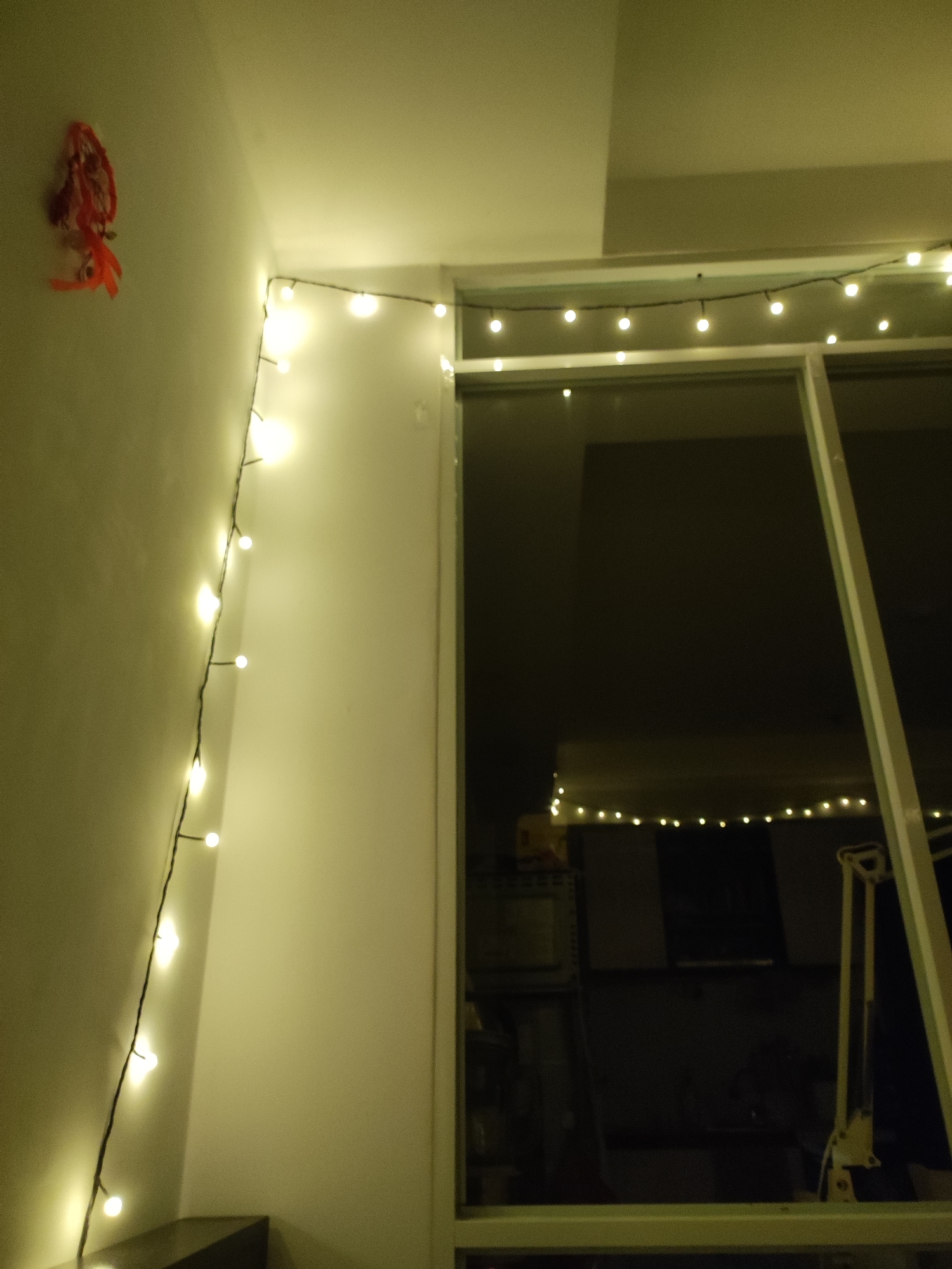 The image shows a close-up, low-angle view of a string of fairy lights attached to a white wall and a window frame. The lights are small, round, and emit a warm, yellow glow. They are draped along the wall and across the top of the window. A red and orange decorative item, possibly a dreamcatcher or a bundle of ribbons, is hanging on the wall to the left of the lights. The window appears to be large and reflective, with the dark interior of a room visible behind it. A tall, articulated lamp is partially visible within the reflected interior. The overall scene suggests an indoor setting, possibly a bedroom or living area, decorated with ambient lighting. The location is specified as Nha Trang Ward, Vietnam, though the image itself does not contain any specific geographical markers to confirm this. The lighting is dim, creating a cozy and festive atmosphere.
