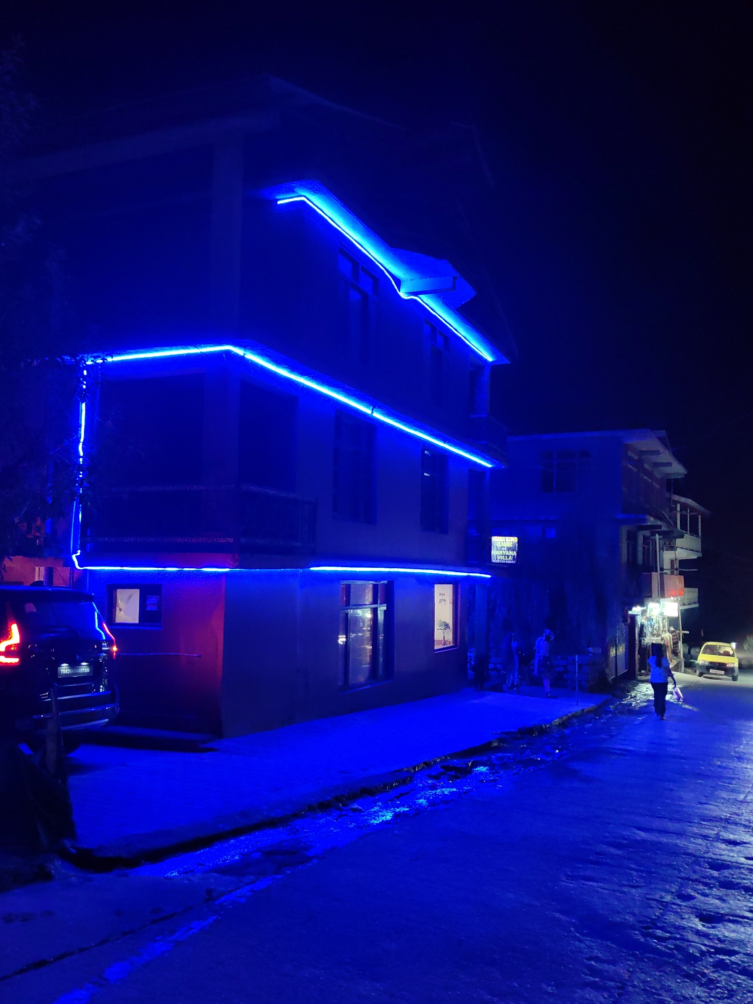 In Barheta, India, a street scene unfolds at night under the vibrant glow of blue neon lights. The primary focus is a building adorned with these luminous strips, outlining its structure and windows. To the left, the rear of a dark-colored SUV is visible, its taillights illuminated. Further down the street, a yellow car is parked, and a person is walking away from the camera, heading towards the car. Other figures can be seen standing or walking in the vicinity of shops lining the street. The road surface appears wet, reflecting the blue light, suggesting recent rain or a damp environment. The overall atmosphere is one of a lively urban night, enhanced by the striking artificial illumination.