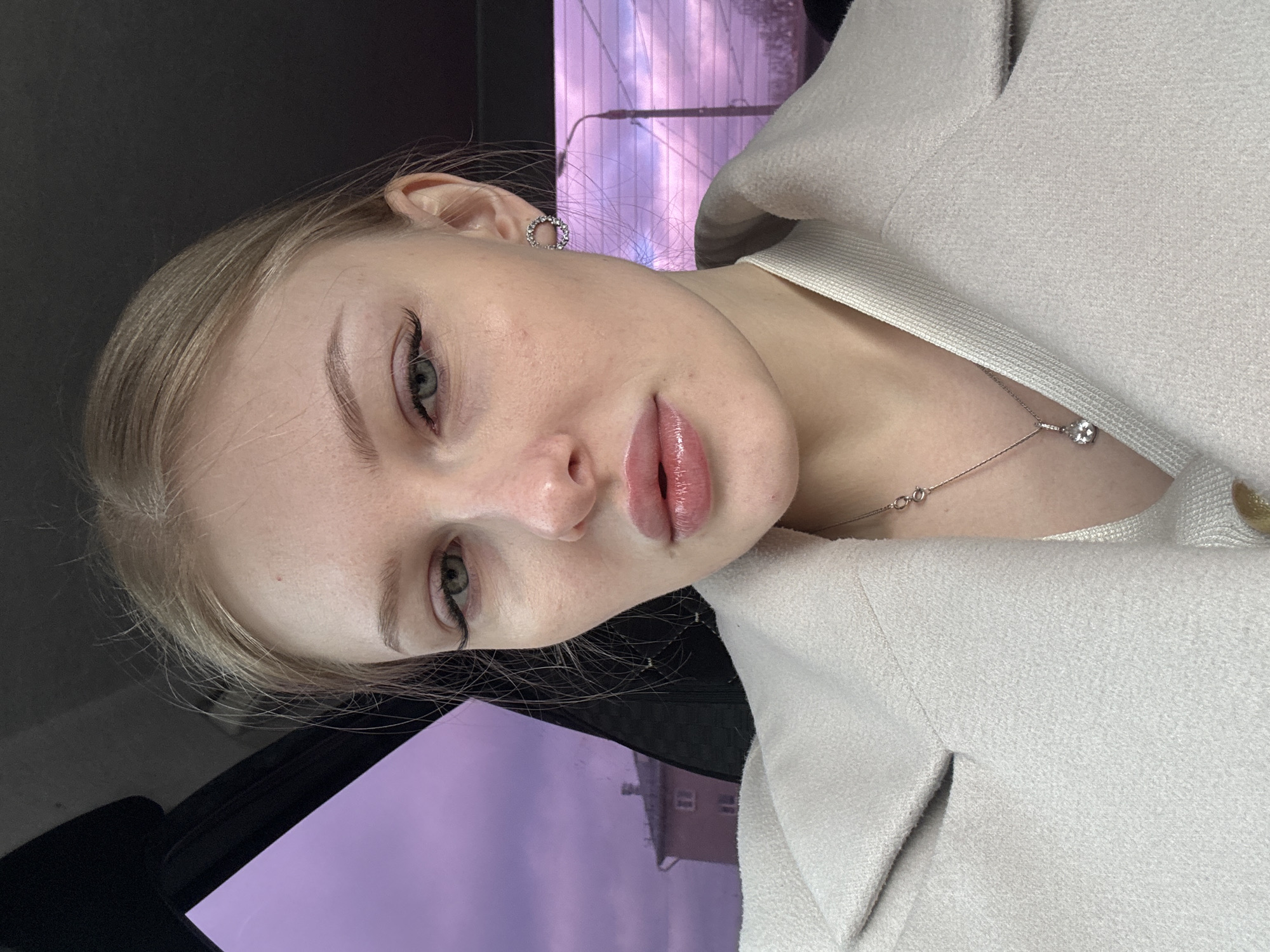 A young woman with fair skin and blonde hair poses for a selfie, wearing a light-colored jacket and a silver necklace. She has light green eyes and is wearing subtle makeup. Her earring is a small, circular design with embedded stones. The background reveals a glimpse of Perm, Russia, through a window, featuring a building with a purplish hue, indicating either a sunset or artificial lighting. The setting appears to be inside a vehicle, with the dark frame of the car window visible in the lower left.