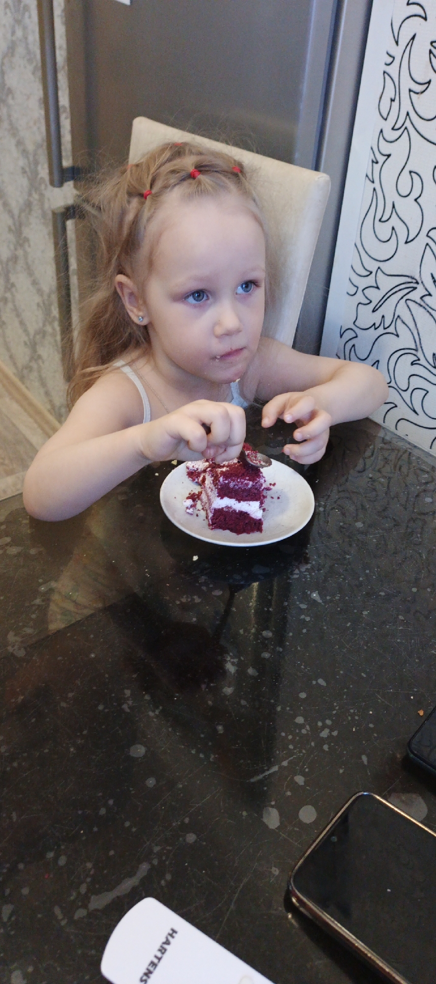 An indoor scene in Chaikovsky, Russia, depicts a young female child, estimated to be between three and five years old, with light skin, blonde hair styled with multiple small red hair ties, and blue eyes. She is seated at a dark, speckled table, occupying a light-colored upholstered chair. The child wears a light-colored, possibly white or light blue, spaghetti strap top. Her right hand holds a silver-colored spoon, which is inserted into a partially consumed slice of red velvet cake on a white ceramic plate. The cake has red layers with white frosting and is lightly dusted with powdered sugar, with crumbs scattered on the plate and table surface. Traces of frosting or crumbs are visible around the child's mouth. Her gaze is directed towards the upper right.
In the background, a silver-colored appliance, likely a refrigerator, with a vertical handle is visible on the left. To the right, a white panel features black decorative scrollwork. Adjacent to the appliance on the left, a wall section with a light-patterned wallpaper is visible. On the table in the lower left foreground, a white object with the partially visible word "HARTENS" is present. A dark-colored smartphone is situated on the table in the lower right foreground.