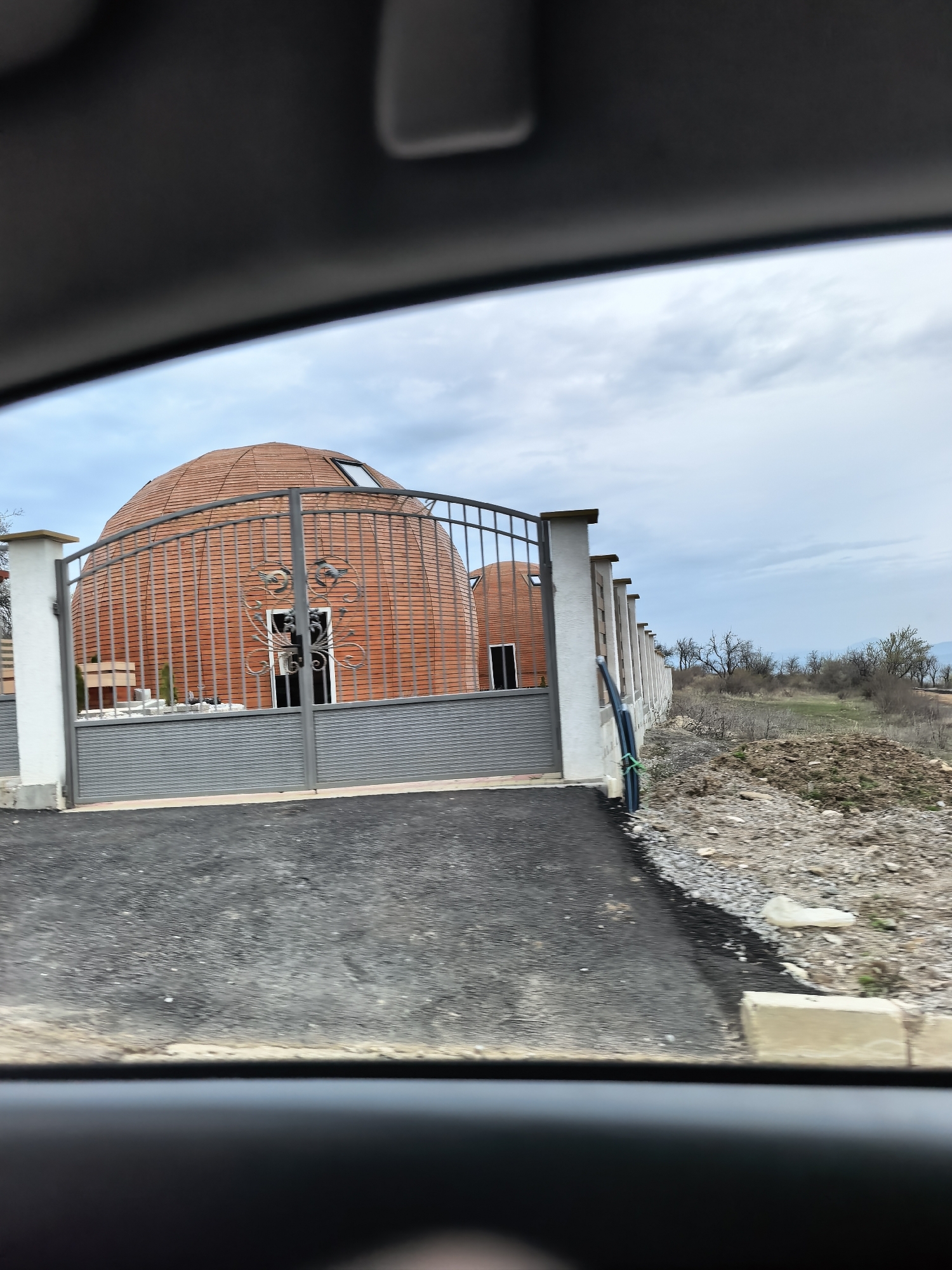 The image shows two rounded, tiled buildings behind a grey metal fence. The buildings have dark-colored doors and what appears to be a skylight on one. The fence is flanked by white pillars. There's a paved road in front of the fence, and beyond the fence is a gravel area. The background shows a field with some trees and an overcast sky. The image is taken from inside a vehicle. The location is Unknown, Unknown.