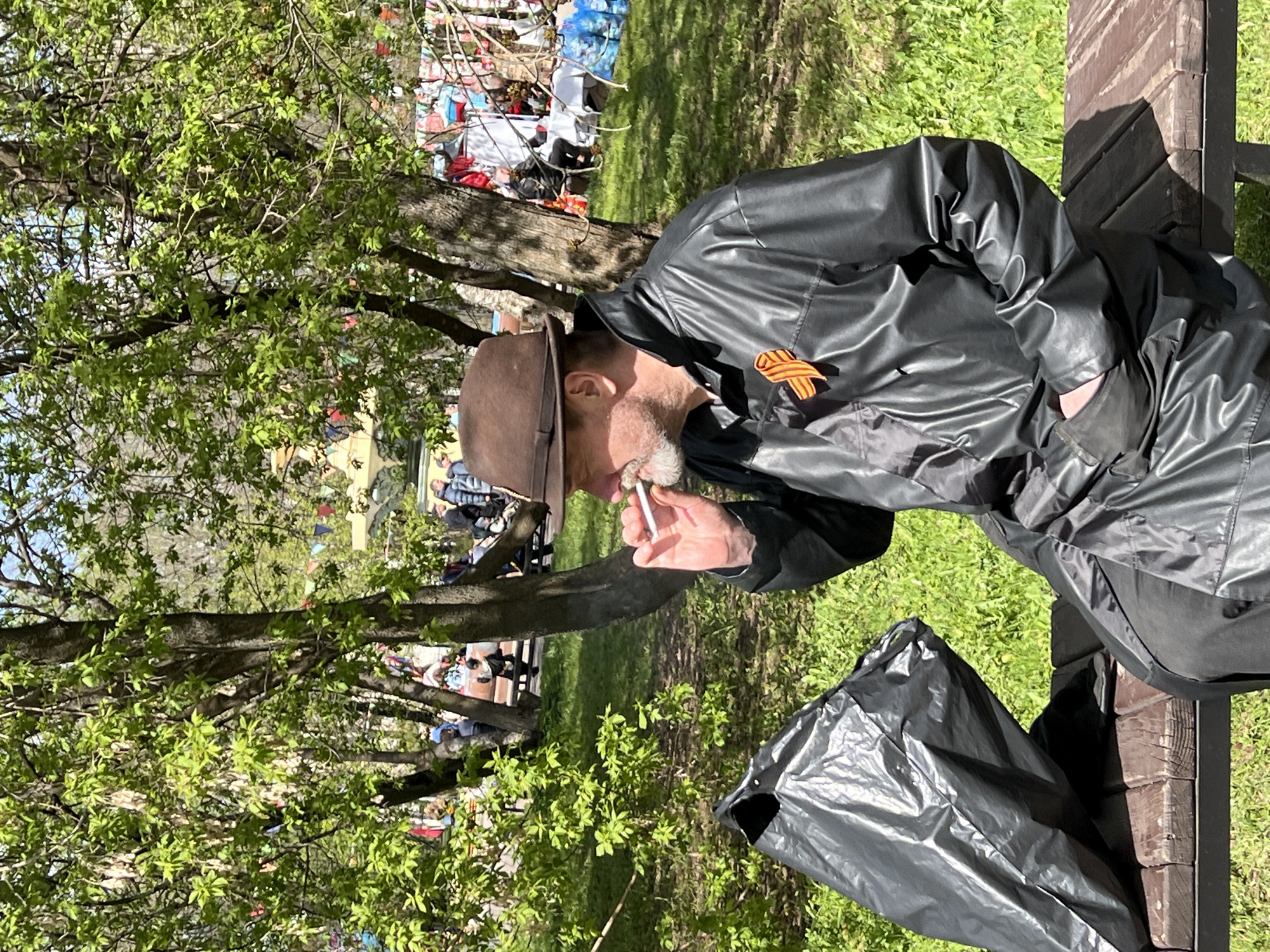 A man with a salt-and-pepper beard, wearing a brown hat and a black, shiny jacket with an orange and black striped ribbon attached, is smoking a cigarette while sitting on a wooden park bench. Next to him on the bench is a black plastic bag. The location is a park with green grass and leafy trees, and there are people visible in the background. This image was taken in Cherepovets, Russia.