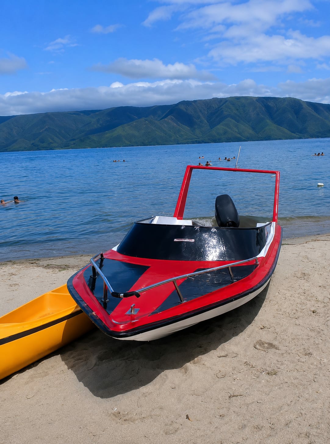 A red and black speedboat is parked on a sandy beach in front of a calm, blue body of water. The boat has a windshield and a roll bar. To the left of the speedboat, a yellow kayak is also pulled up onto the beach. In the background, lush green mountains rise from the water under a bright blue sky with scattered white clouds. Several people are visible swimming and wading in the water, with more in the distance. The location is Situngkir, Indonesia.