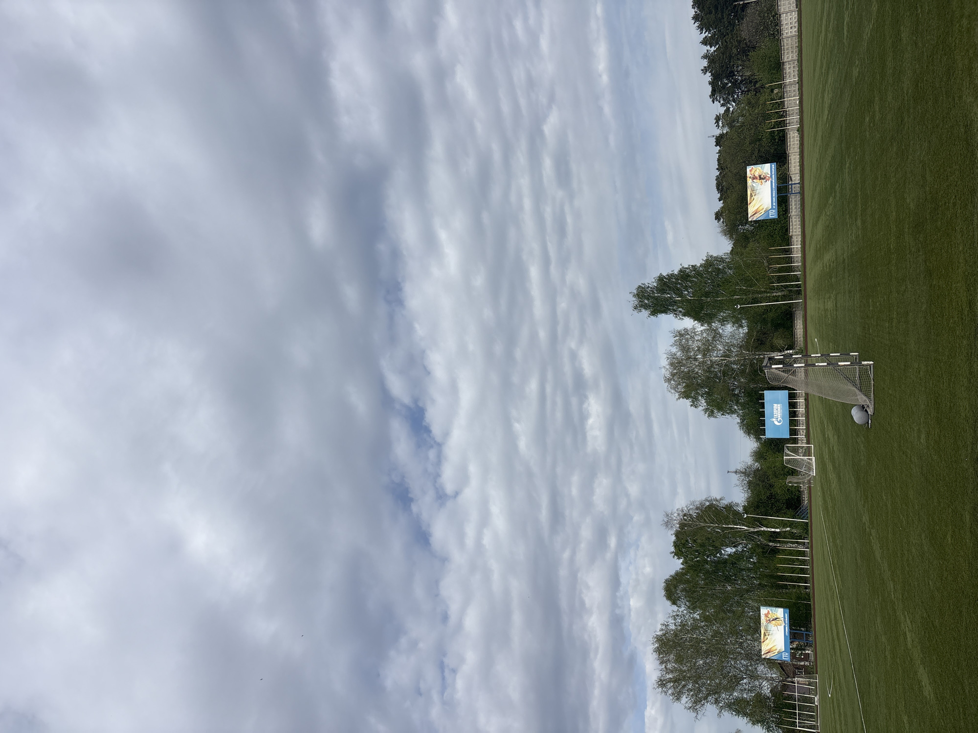 The image shows a sports field in Chaikovsky, Russia. The sky is mostly cloudy with some patches of blue. The field has green artificial turf and is lined with a fence and trees. There are three advertising screens along the edge of the field. Two soccer goals are visible, one with a blue Gazprom banner. A soccer ball is visible near the goal.