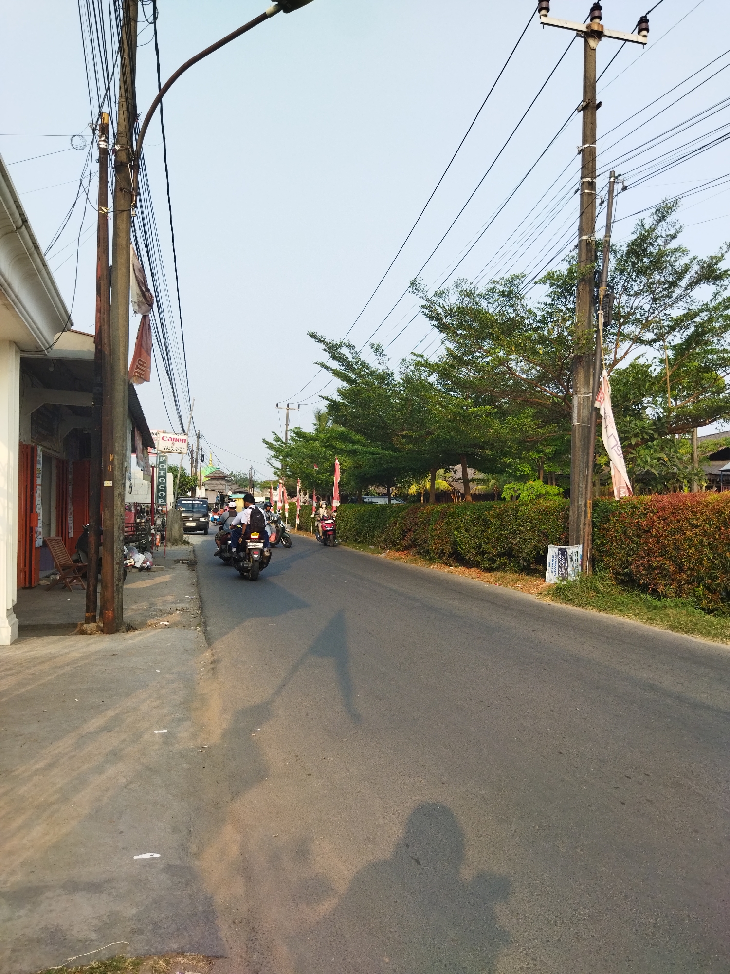 The image depicts a street scene in Suka Sari, Indonesia, under a clear sky. On the left side of the asphalt road is a sidewalk with a building that appears to be a shop, with its shutters partially closed. In front of the shop, there are two chairs and a wooden table. A lamppost stands on the sidewalk.
The road itself has several motorcycles on it. One motorcycle in the foreground has two people riding it, one driving and one passenger wearing a white shirt and a black backpack. Another motorcycle is visible further down the road. There is a black SUV driving in the same direction as the motorcycles.
The right side of the road is lined with a dense, well-maintained hedge with red and green foliage. Behind the hedge are several trees with lush green leaves and a tall wooden utility pole with numerous electrical wires. A flag is attached to the utility pole. Further down the road, more trees and what appears to be a building with a green roof are visible.
The time of day appears to be late afternoon or early evening, as indicated by the long shadows cast on the road and sidewalk. The overall impression is of a moderately busy street in a residential or commercial area.