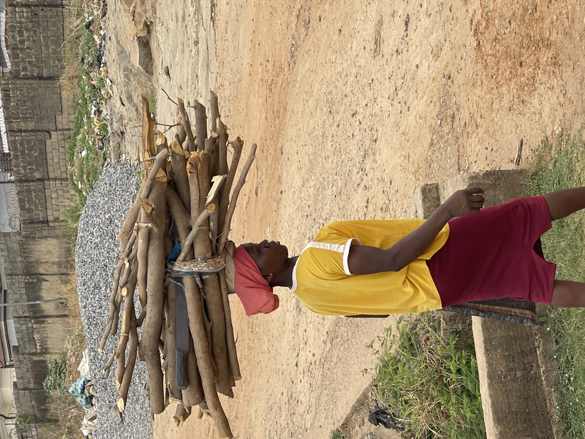 A woman is carrying a bundle of firewood on her head in Abuja, Nigeria. She is wearing a yellow shirt with white trim and a red skirt. A red headscarf covers her hair. The firewood is a large pile of thin, brown branches bound together with a patterned cloth. A large, black machete is nestled in the bundle. The woman is standing on a dirt path with some grass growing nearby. In the background, there is a pile of gray gravel, a concrete wall, and some scattered trash.