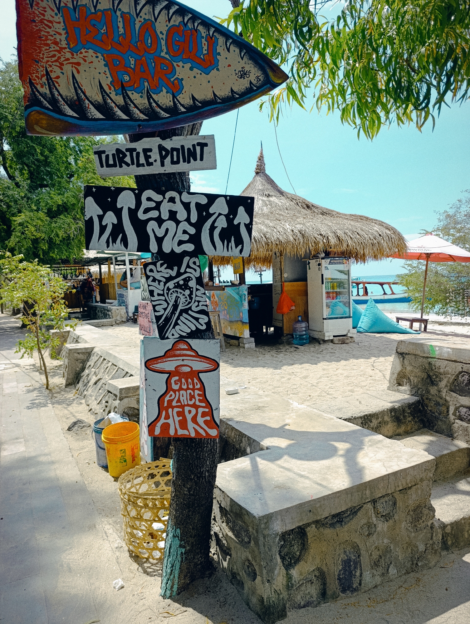 This image depicts a vibrant street scene in Gili Trawangan, Unknown, Indonesia. A weathered wooden pole displays multiple hand-painted signs: "HELLO GILI BAR", "TURTLE POINT", "EAT ME", a psychedelic mushroom graphic, and a red UFO illustration with the text "GOOD PLACE HERE". The pole is surrounded by a woven basket and a yellow bucket.
In the background, a thatched-roof structure with a beverage refrigerator stands on a sandy area, suggesting a beachfront establishment. A blue beanbag chair and a table are visible near an umbrella. The sea is in the distance with a boat floating on the water. The scene is captured during the day under a clear, sunny sky with trees lining the street. The sidewalk consists of concrete structures.