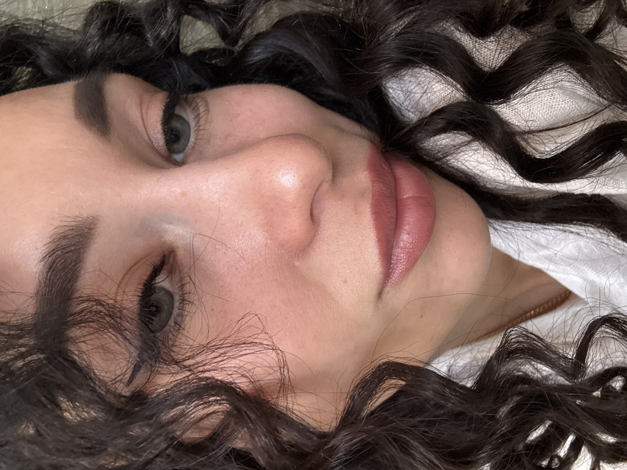 A close-up portrait of a woman with curly, dark brown hair. She has light skin, dark eyebrows, and green eyes. Her lips are a muted pink color. She is lying on a white fabric surface. There is no evidence to suggest the location is Chaikovsky, Russia.