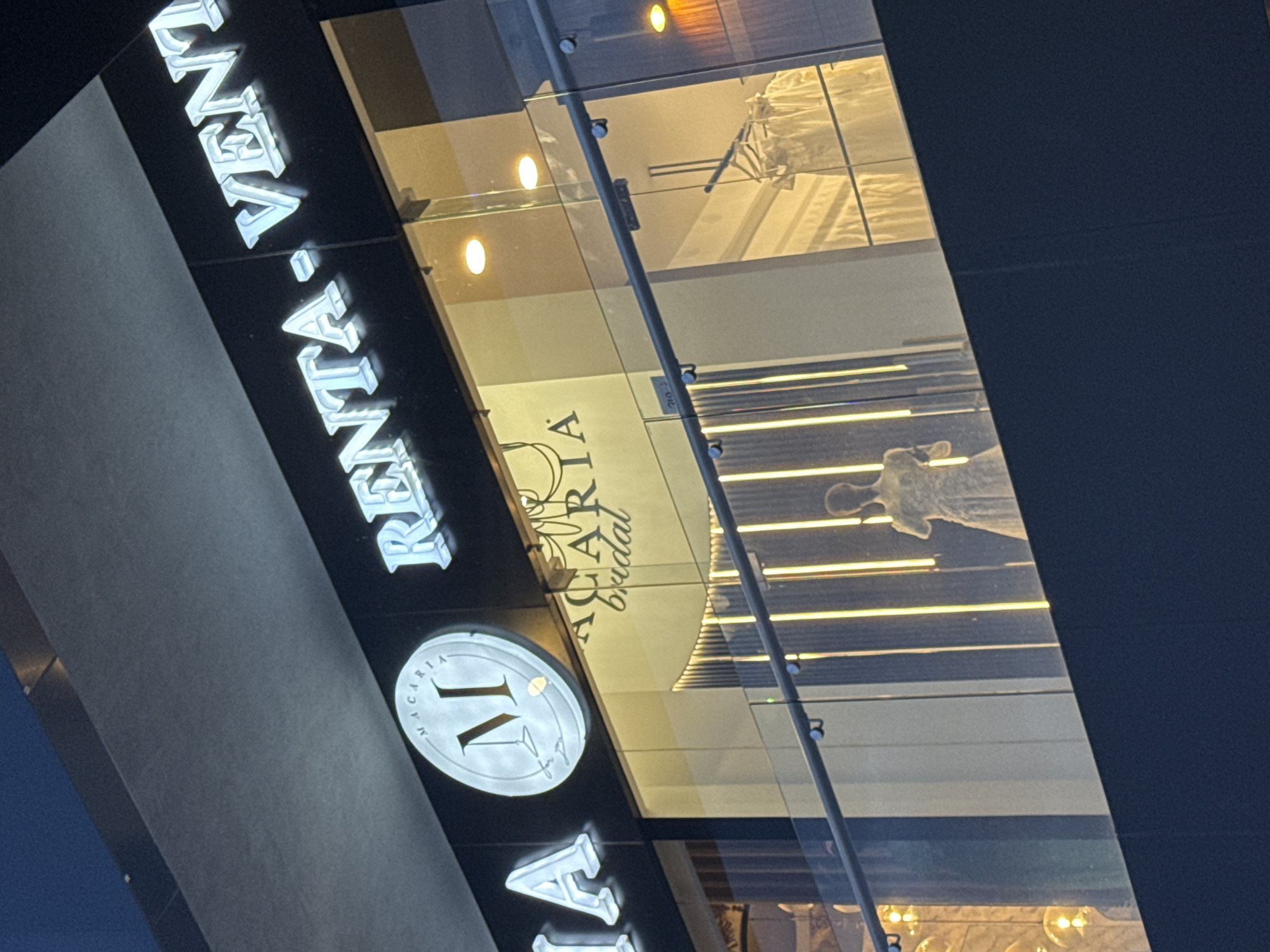 This image captures the exterior of a modern commercial building at night or dusk in San Bernardino Tlaxcalancingo, Mexico. The scene is dominated by a large, illuminated sign on the building's facade, partially reading "RENTA VEN", possibly "RENTAS VENUES" or similar. Below this sign, there is a prominent circular illuminated emblem featuring a large, stylized letter "M" with the word "MACARIA" arcing above it. Beneath the 'M', subtle graphics of martini glasses and the text "By Gigi" are visible, suggesting a sophisticated establishment.
The building features a multi-pane glass facade that reflects the interior lighting. Through the glass, distorted but legible text reads "MACARIA Bridal," indicating the business is likely a bridal shop or a venue specializing in weddings. The interior visible through the glass is brightly lit with warm, yellow-toned lights, including multiple oval or circular fixtures and horizontal strip lighting. A large white object, possibly a wedding dress on a mannequin or a decorative element, is faintly discernible behind the glass in the upper right section. The glass panels are secured with visible bolts and metal framing, and a small 'B' is marked on some of the structural elements. The overall impression is one of a stylish and upscale commercial venue.