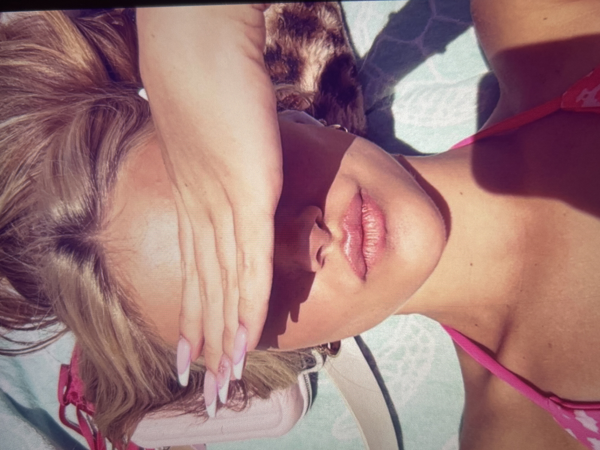 This is a close-up, overhead, point-of-view shot of a person's face partially covered by their hand. The subject has blonde hair and is wearing a pink bikini top. The background appears to be a blue and white patterned towel or blanket. There is a shadow cast across the person's face, suggesting they are outdoors in sunlight. The image does not provide any information about the city of Knezha, Bulgaria, or any other geographical location.
