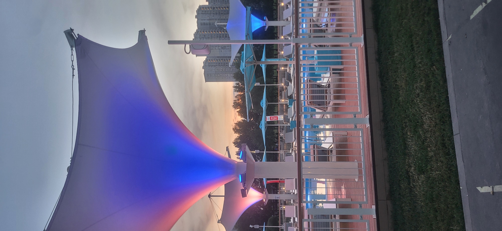 A poolside lounge area is depicted at dusk. Several lounge chairs with white frames and grey cushions are arranged around a pool. White railings with vertical slats separate the deck from the surrounding grassy area. Large, white tensile fabric structures, illuminated with colorful blue and pink lights, provide shade. In the background, a line of trees separates the lounge area from several multi-story residential buildings. The sky is overcast, suggesting either the end of a day or approaching weather. There are no people visible in the image.