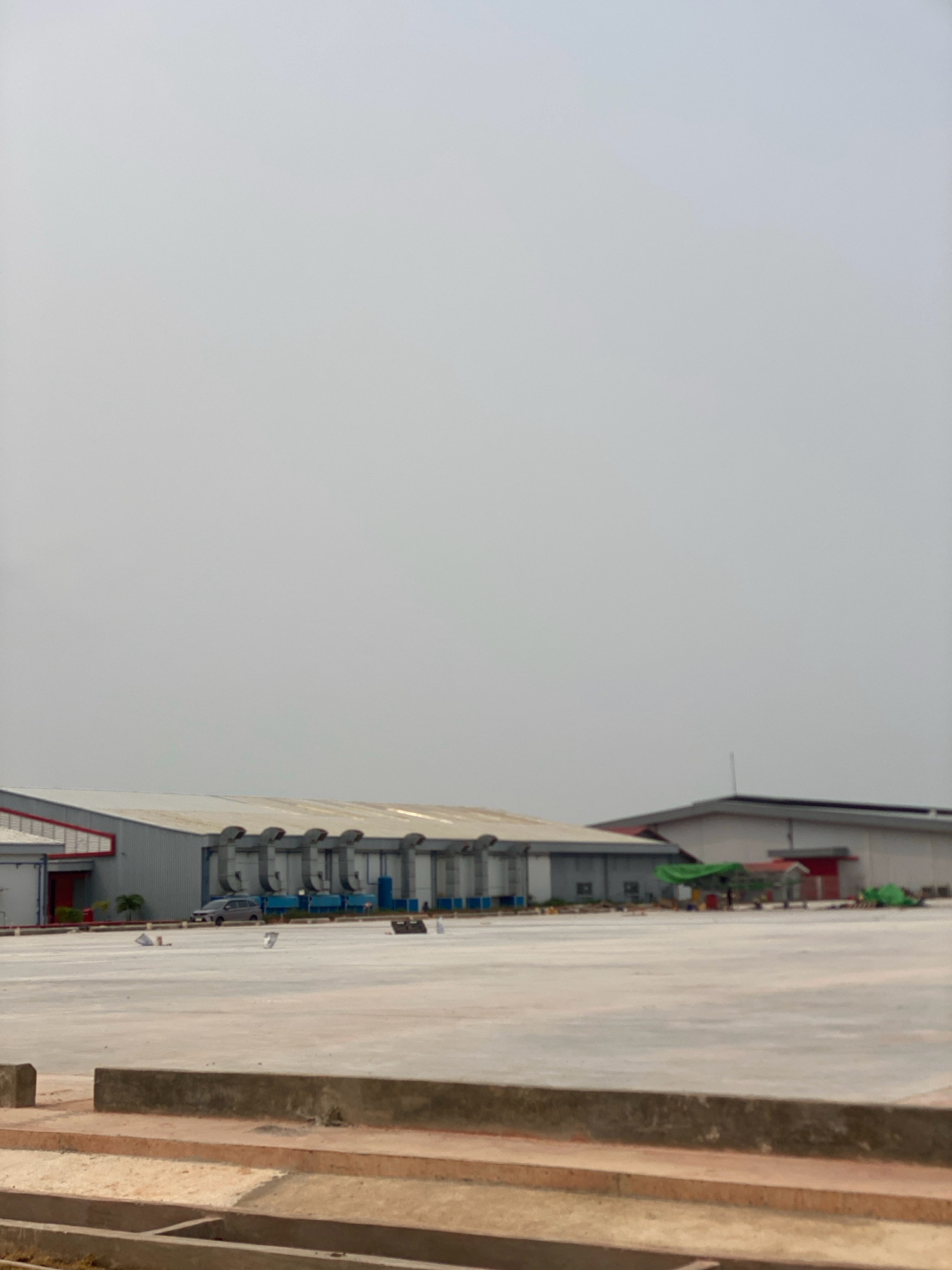 The image shows a large, flat, paved outdoor area, likely a parking lot or staging area, in front of industrial buildings. The buildings are utilitarian in design, with metal siding and large roll-up doors. One building has a series of large, industrial-looking ventilation units along its roofline. There is a silver car parked in front of one of the buildings. In the foreground, there are concrete structures, possibly curbs or retaining walls. The sky is overcast and a hazy gray. The scene appears to be located in an industrial zone in Batam, Indonesia, likely within a logistics or manufacturing facility. There are no people visible in the image.