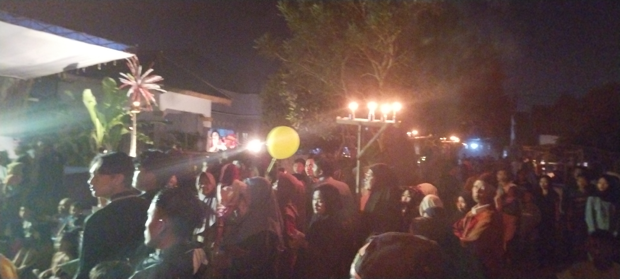 The image depicts an outdoor gathering at night, possibly in the city of Keting, Indonesia, indicated by the prompt. A large crowd of people is visible, many of whom appear to be observing an event or performance taking place under a white canopy on the left side of the frame. Several individuals are in the foreground, looking towards the left, with some wearing dark clothing.
To the right of the canopy, a tall pole with several lit candles or lamps stands as a focal point, casting a warm glow. A bright yellow balloon is also visible in the mid-ground, held by an unseen person. In the background, there are trees and structures, suggesting a semi-urban or rural setting. The lighting is dim, with artificial lights from the candles and possibly other sources illuminating the scene, creating a festive or ceremonial atmosphere. The overall impression is of a community event happening after dark.