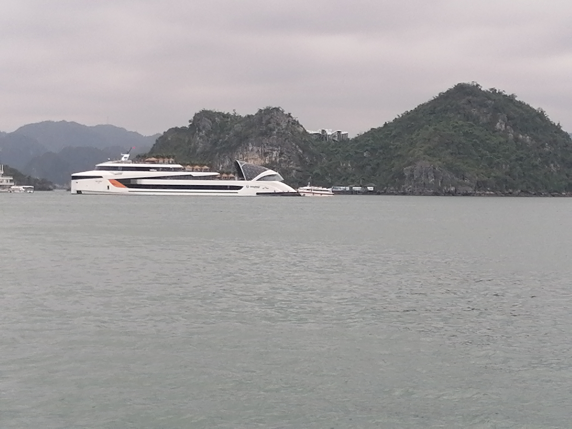The image captures a serene waterscape dominated by towering limestone karsts under an overcast sky. In the mid-ground, a large, modern, white catamaran-style vessel, identified by the text "Ambassador CRUISE" on its hull, takes center stage. This sleek passenger boat features multiple decks, a distinctive orange and black stripe near the bow, and a modern, curved superstructure towards the stern. A flag is visible flying from its mast.
The boat appears to be stationary, possibly docked or anchored, amidst the calm, grayish-green water, which shows gentle ripples. Behind the main vessel, a smaller white boat is partially visible, and further to the right, another smaller boat is moored near the shore of an island.
The background is filled with the iconic, lush green limestone islands of Ha Long Bay, Vietnam, consistent with the provided location context. These karsts are steep and covered with dense vegetation, with some exposed rock faces. On one of the larger islands, several contemporary buildings or structures are subtly visible amidst the trees, suggesting development or resorts integrated into the natural landscape.
The weather is cloudy and muted, creating a soft, diffused light across the scene. The overall color palette is subdued, with grays, greens, and whites. No human figures are distinctly visible, and the scene conveys a tranquil atmosphere, likely depicting a moment during a tour or journey through the bay.
