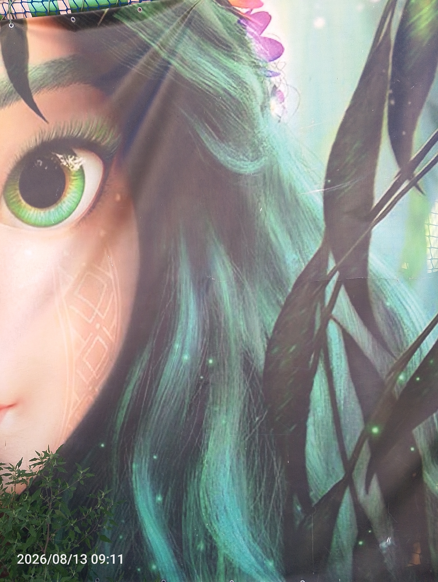The image is a close-up of a mural or advertisement depicting a young woman with striking green eyes and long, flowing green hair. She is adorned with a floral headdress, and her face features intricate, glowing patterns. The background is a soft, ethereal blue-green, with hints of darker foliage. In the lower left corner of the image, a small, leafy plant grows in front of the mural. The bottom edge of the image displays a timestamp: "2026/08/13 09:11". The mural is located in the city of Bahacheve, Ukraine.