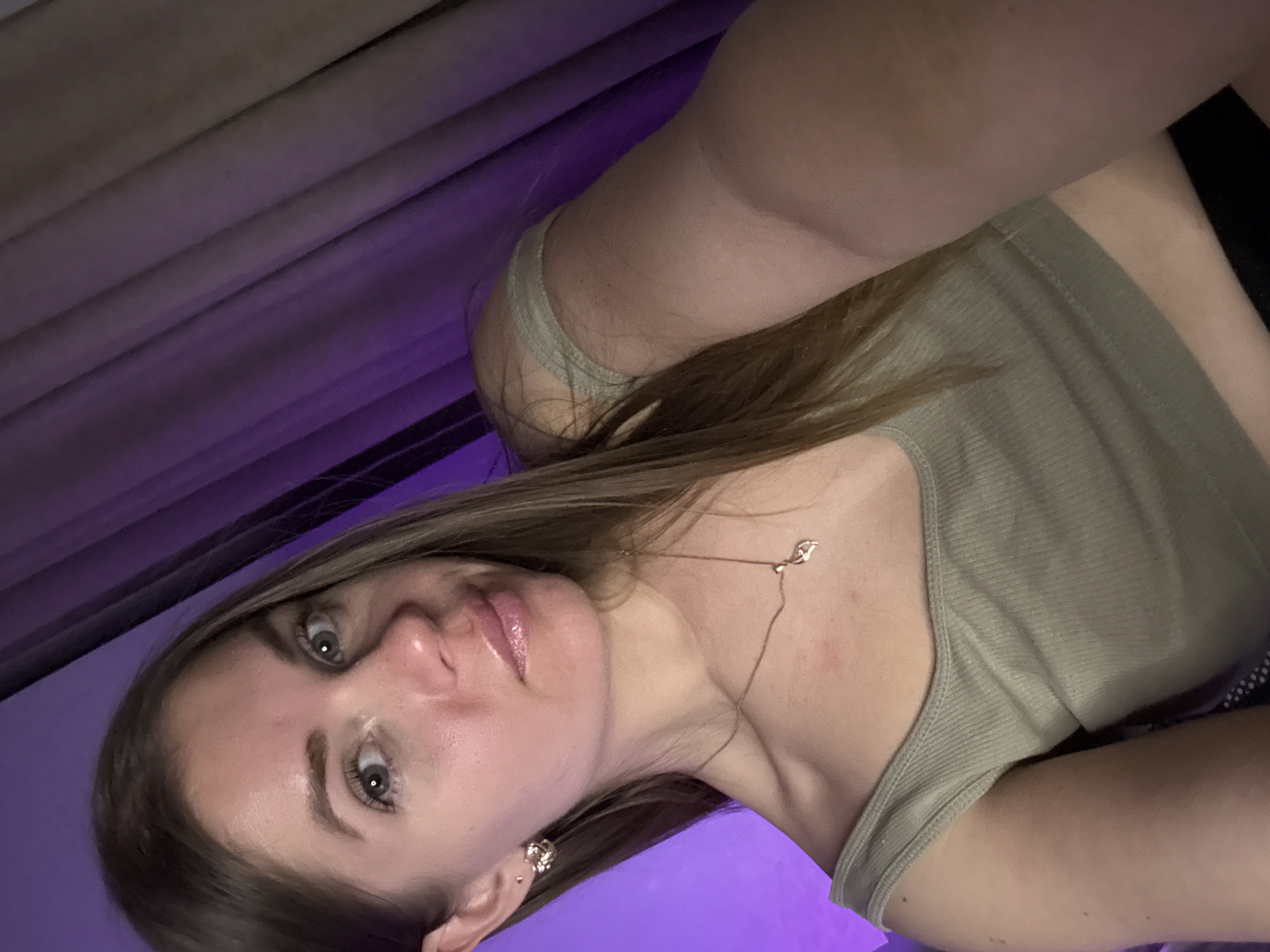 A selfie photograph shows a light-skinned woman with long brown hair wearing a light green tank top and jewelry. The woman has gray eyes and glossy lips. The background consists of beige curtains and a purple light source. The location is not discernible from the image.