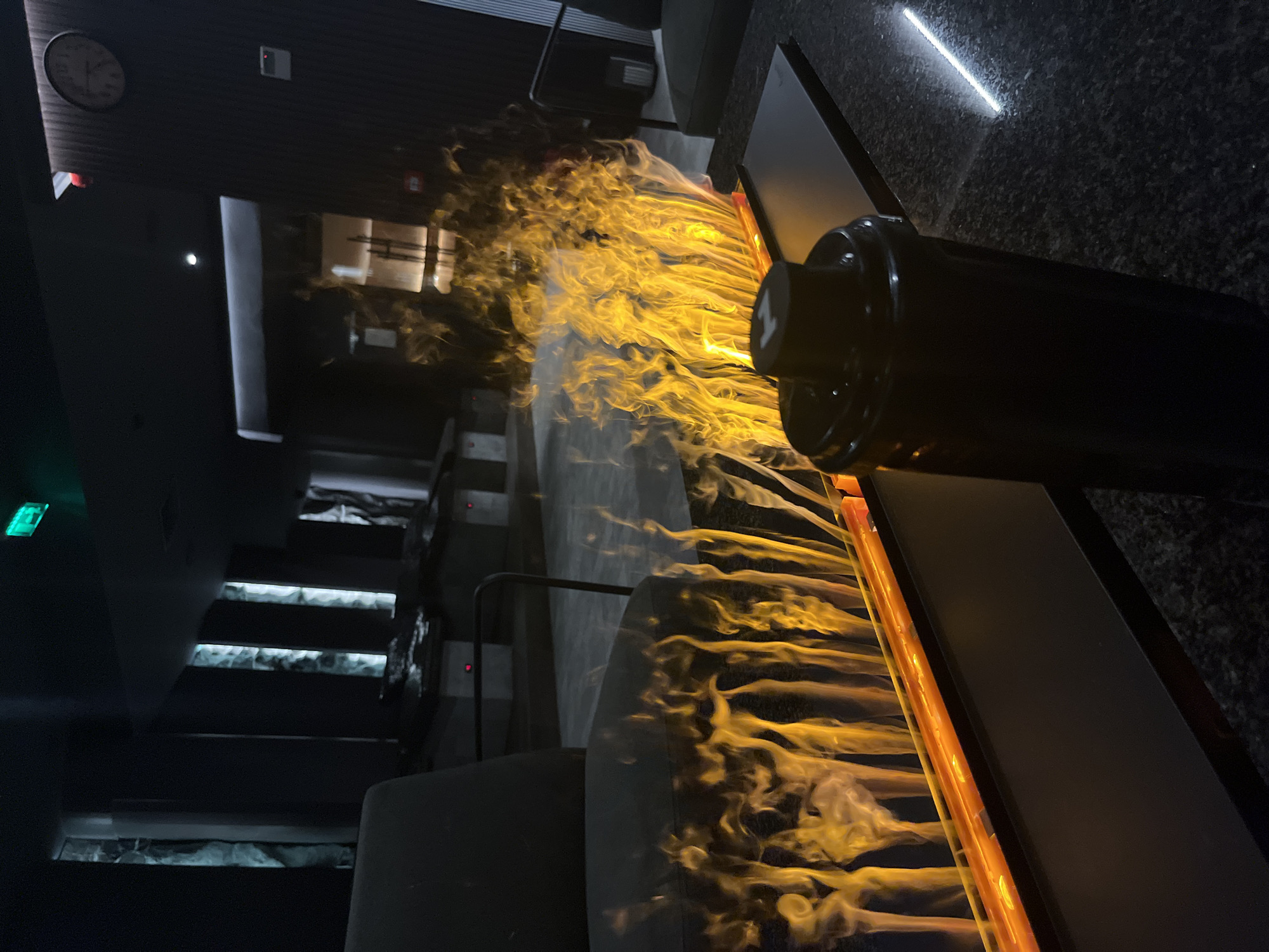 The image depicts an interior space in Sofia, Bulgaria, featuring a decorative element: a long, thin trough filled with what appears to be simulated flames or illuminated mist, creating a fire-like visual effect. To the right of the trough, a black, cylindrical object (possibly a vase or decorative piece) is situated on a dark, granite-like surface. The overall lighting is dim, enhancing the contrast between the bright "flames" and the surrounding dark environment. In the background, a set of stairs with embedded lights can be seen, and further back, what appears to be a seating area with multiple chairs. A clock is visible on the wall in the background. The perspective is from a low angle, possibly at table height.