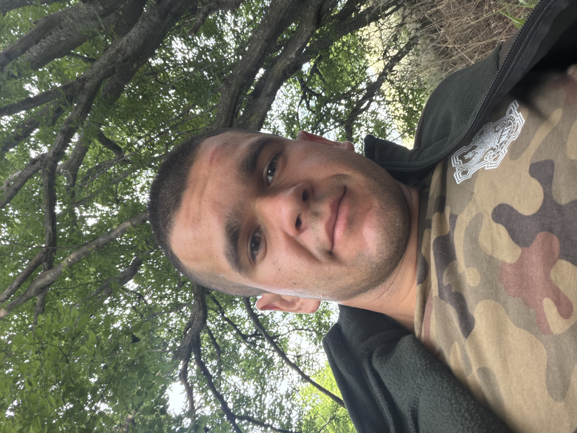 This is a medium shot selfie taken outdoors, with the camera angled upwards towards a person and a tree. The person, a man with short dark hair and a light complexion, is positioned in the lower half of the frame, looking towards the camera. He is wearing a camouflage t-shirt with a white emblem on the left chest, and a dark green zippered jacket over it. The man's face shows a slight smile.
The background is dominated by a large tree with thick branches and lush green leaves, filling the upper portion of the image. The sunlight filters through the leaves, creating a dappled effect. Beyond the tree, a hazy, dry vegetation and the sky are visible. The overall impression is of a casual outdoor setting, possibly a park or a wooded area.
The lighting suggests it is daytime, likely morning or afternoon, with no strong shadows indicating a specific time. The presence of green leaves suggests it is spring or summer. The man's attire is casual outdoor wear. No specific activity is clearly depicted, but the selfie angle suggests the person is taking the photo themselves while enjoying the outdoors. There is no visible text in the image. The location context provided is Ledenik, Bulgaria, which aligns with the natural outdoor scenery.