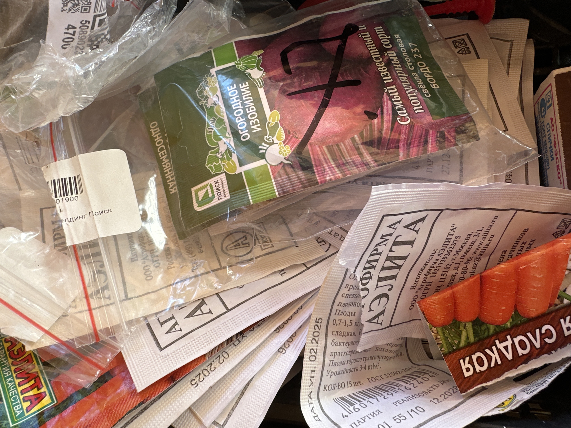 This close-up image captures a collection of various seed packets and small transparent plastic bags, creating a cluttered scene likely inside a storage container or box. The primary subjects are seed packets for vegetables, predominantly written in Russian, indicating a context within Russia, such as Nizhny Novgorod.
The main activity suggested is the storage or organization of gardening supplies, likely in preparation for planting or for safekeeping after harvesting. No specific event is actively taking place; rather, the image shows items at rest.
Notable details include the diverse packaging designs of the seed packets. One prominent packet features an illustration of a beetroot, with text identifying it as "БОРДО 237" (BORDO 237) table beet, from "ПОИСК Агрохолдинг" (POISK Agroholding), described as "Самый известный и популярный сорт" (The most famous and popular variety). Another packet, partially visible, displays an image of carrots and prominently features the brand "АЭЛИТА" (AELITA), with details such as "РЕАЛИЗОВАТЬ ДО 12.2028" (SELL BY 12.2028) and "ДАТА УП. 02.2025" (PACKING DATE 02.2025). This packet also specifies a quantity of "15 шт." (15 pcs.) and references "ГОСТ 32592-2013" (State Standard 32592-2013), and details like "сладкая" (sweet) and "Плоды 0,7-1,5 кг" (Fruits 0.7-1.5 kg).
Among the seed packets, small clear plastic bags with red zip closures are visible, possibly containing smaller quantities of seeds. A small matchbox, branded "ФЭСКО" (FESKO) and labeled "СПИЧКИ" (MATCHES), is also present, adding to the assortment of utilitarian items. Other visible text fragments include barcodes, numerical codes like "5089023", "4700", "01900", and partial phrases such as "ГАРАНТИЯ КАЧЕСТВА" (QUALITY GUARANTEE). The items appear well-preserved, suggesting they are in active use or being stored for future use. The lighting indicates daylight, but no specific time of day or weather conditions can be discerned. There are no people visible in the image, and no specific emotions are conveyed.