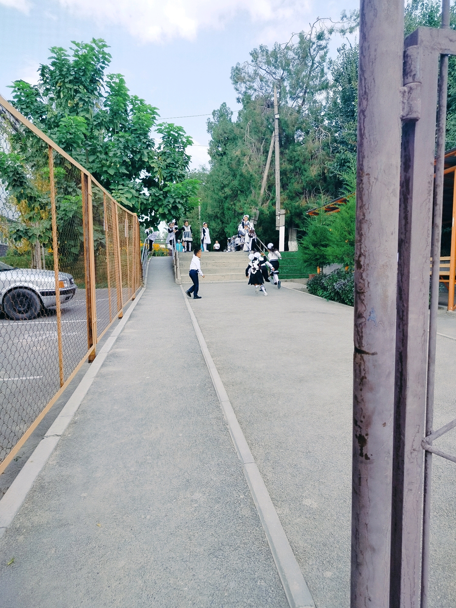 The image depicts a schoolyard scene in Kolkent, Kazakhstan. A silver sedan is parked on the left, behind a chain-link fence with wooden posts. A paved pathway runs parallel to the fence, leading towards a ramp with stairs on the right. Several children in school uniforms are present. Some are standing at the top of the ramp, while others are in motion. One boy in a white shirt and dark pants is walking on the pathway towards the foreground. Further up the ramp, a group of girls in black and white school uniforms are running. The environment is characterized by trees, foliage, and a bright sky with scattered clouds. A large metal gate is visible in the right foreground.