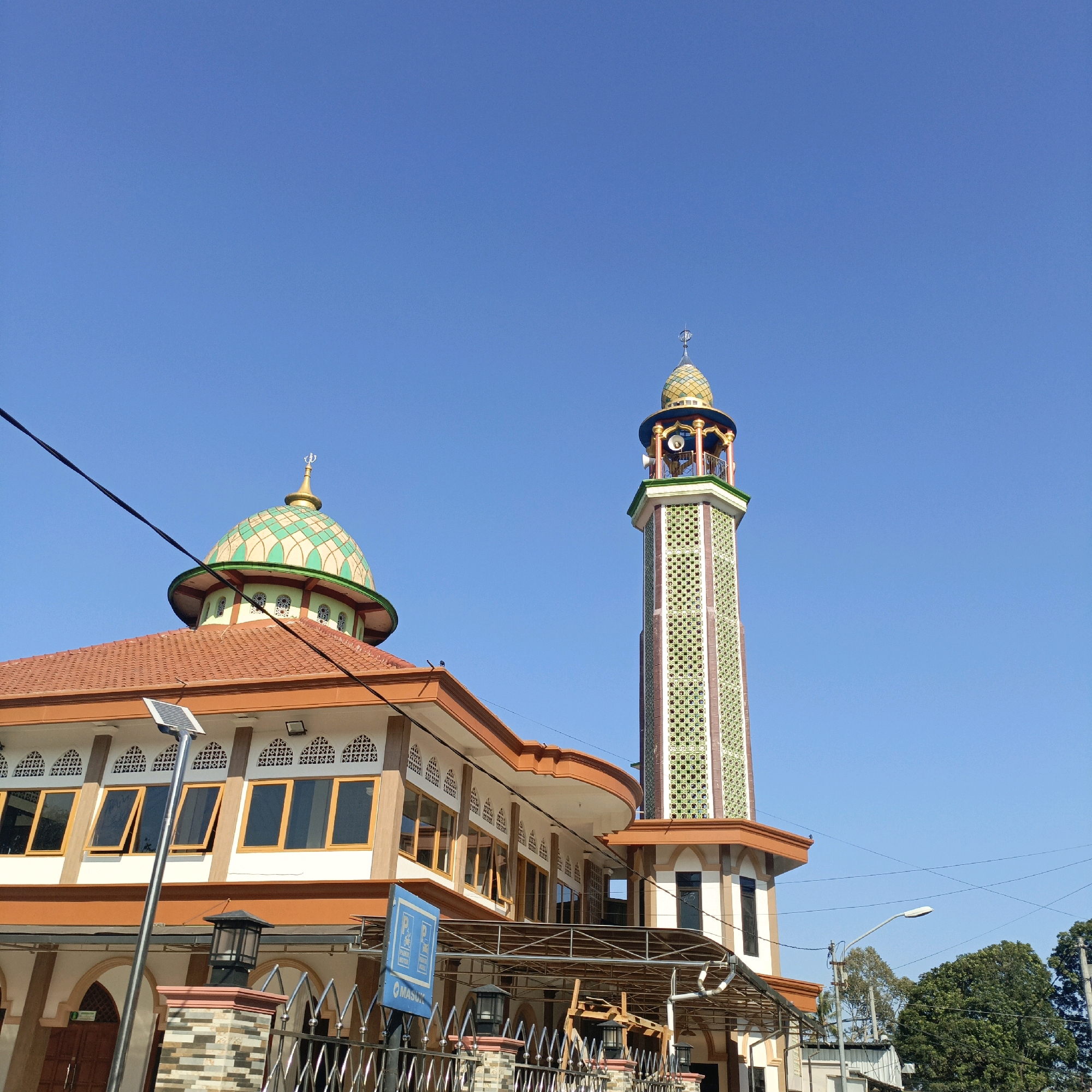 A mosque with an orange tiled roof and a green and gold dome stands against a clear blue sky. A tall, slender minaret, also decorated with green patterns, rises to the right of the main building. The mosque's facade features white walls with decorative windows and brown trim. In front of the mosque, a metal fence encloses the property, with decorative pillars and outdoor lights. A blue sign with parking symbols is visible near the fence. A solar panel is mounted on a pole in front of the building, and power lines are strung across the sky. Trees and some buildings are visible in the background. The location is identified as Salatiga, Indonesia.