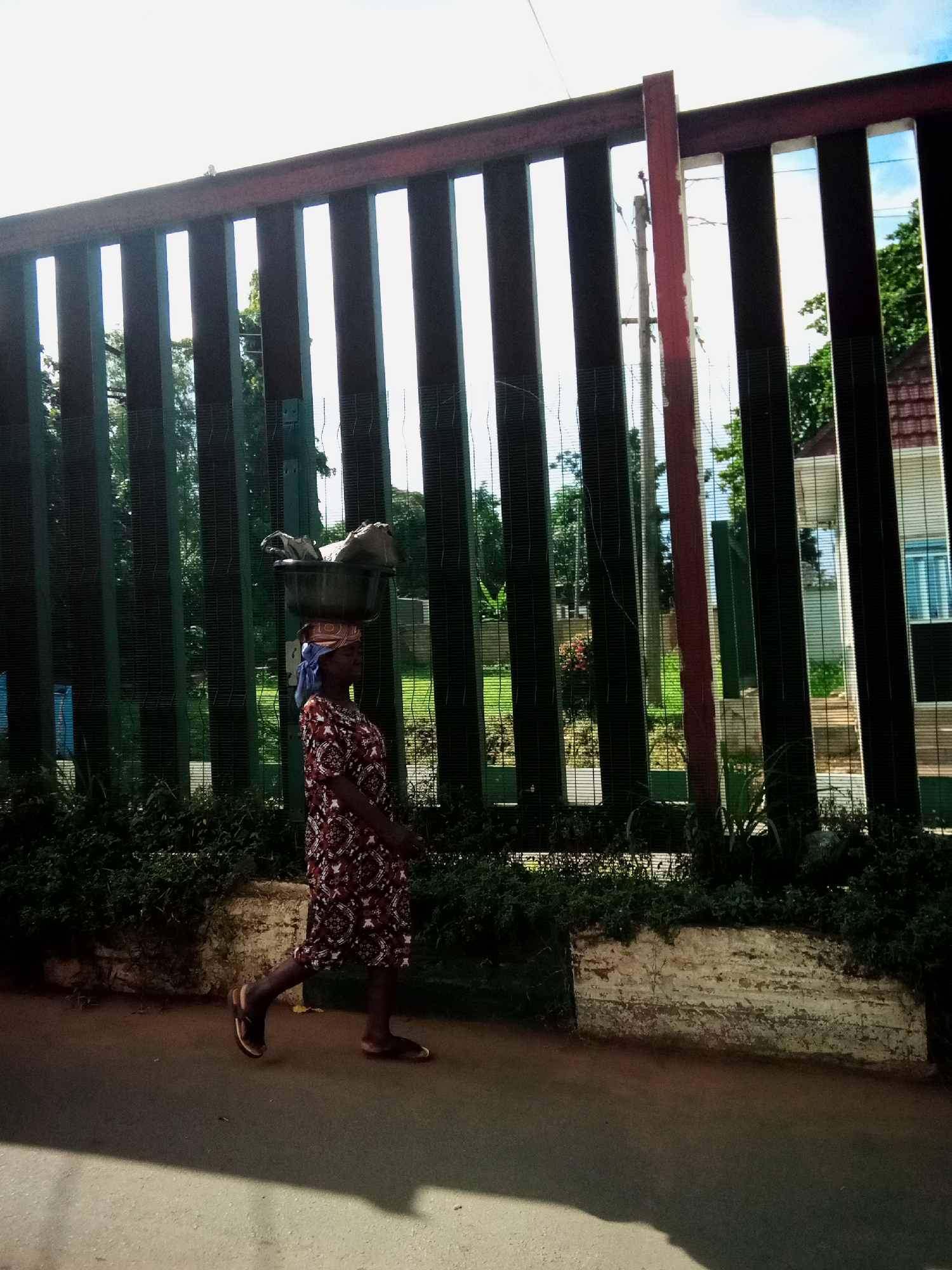 A woman, seen from the side, walks along a paved path in Nibo, Nigeria. She is wearing a patterned dress and a blue headscarf. On her head, she balances a dark-colored bowl filled with various items, including what appears to be fabric. Behind her, a tall, green metal fence with vertical bars runs horizontally across the image. Beyond the fence, trees and parts of buildings are visible. The ground in the foreground is a light brown asphalt.
