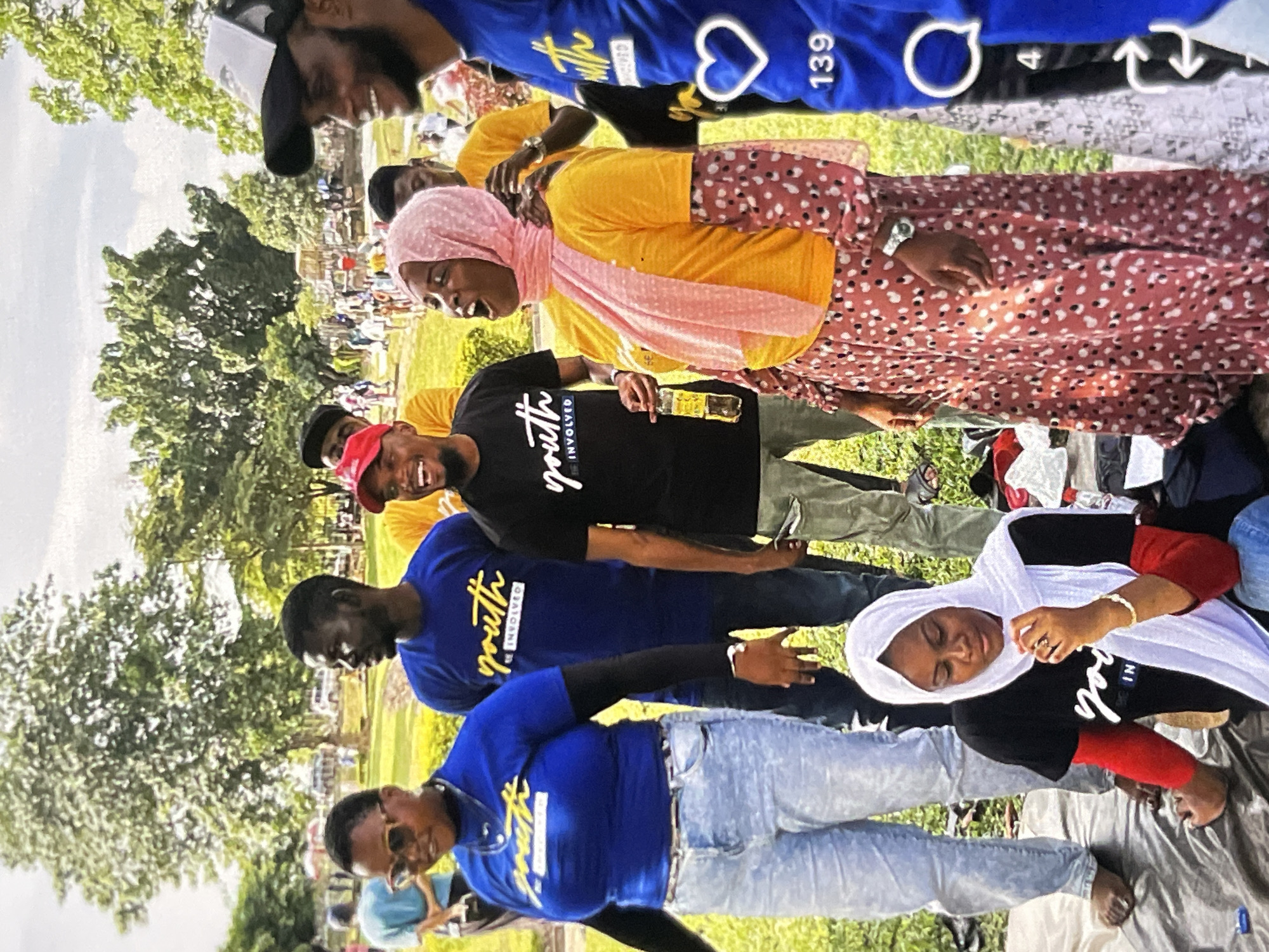 In an outdoor setting, likely a park, a group of young adults are gathered. The overall atmosphere appears casual and social, with individuals in various states of engagement. The background consists of trees, a cloudy sky, and possibly distant structures or vehicles. Several people are wearing t-shirts with the text "Youth Involved" printed on them in yellow lettering.
In the foreground on the left, a woman wearing sunglasses, a blue t-shirt, and light-wash jeans is looking to the side. Next to her, a man in a blue "Youth Involved" shirt is also looking away. To their right, another man in a black "Youth Involved" shirt with a red baseball cap is laughing, holding a bottle. In the center, a woman in a pink hijab and a yellow t-shirt with a patterned skirt is looking upwards, her mouth open. Behind them, other individuals can be seen in the background, some wearing similar "Youth Involved" shirts. To the far right, partially visible, is a person in a blue shirt with social media icons overlayed on the image, showing a heart and the number "139". In the lower portion of the image, a woman wearing a white hijab and a black shirt with red sleeves is seated, looking down. The ground is covered with a dark mat and various items, suggesting a picnic or gathering.