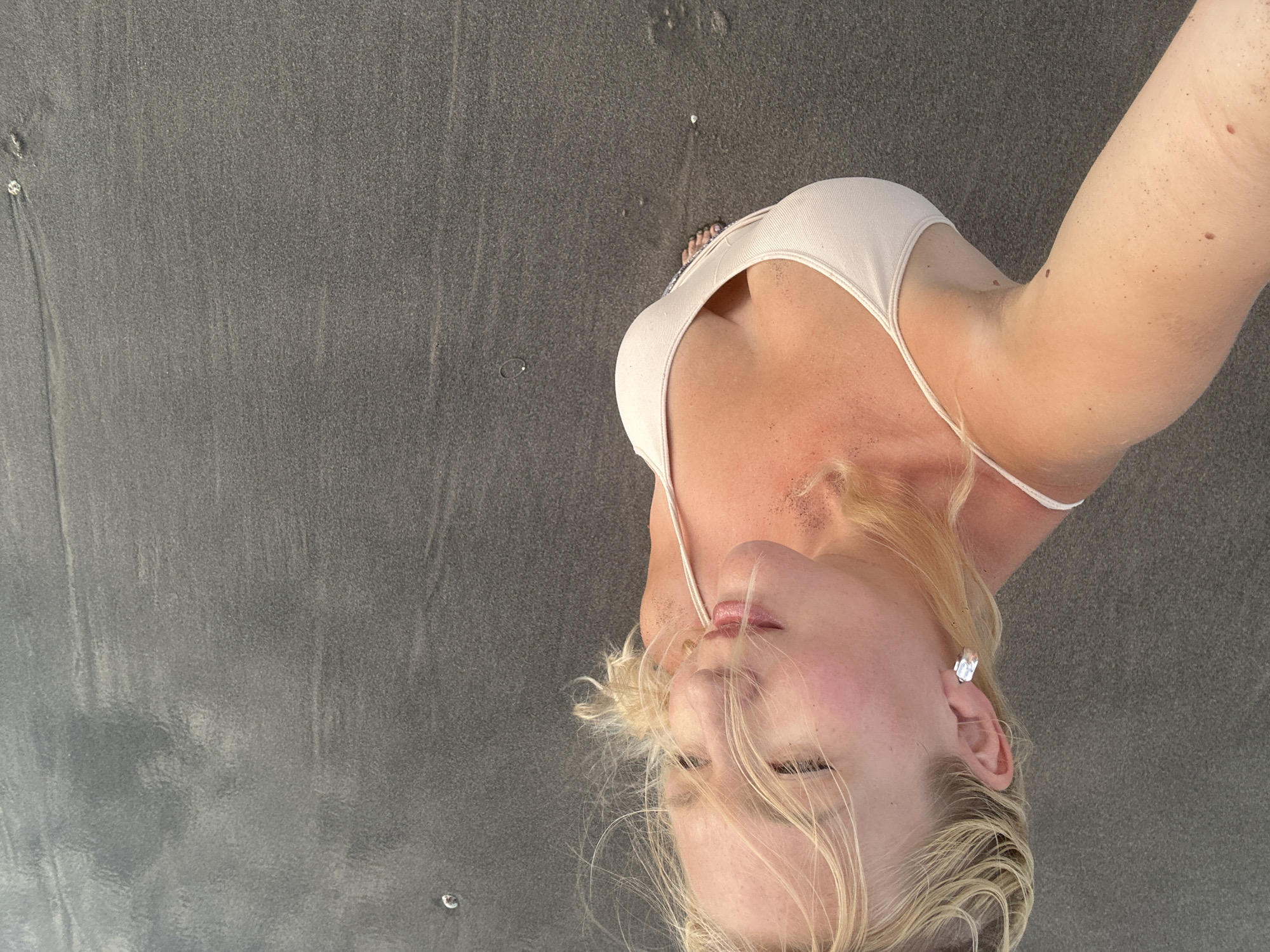 A blonde woman stands on a beach in North Kuta, Indonesia. She is wearing a light-colored bikini top and a silver earring. Her hair is windswept, and her eyes are closed. The sand is dark and wet, with some ripples indicating the tide has recently receded. The lighting suggests it is either early morning or late afternoon. The image is taken from a high angle, looking down at the woman.