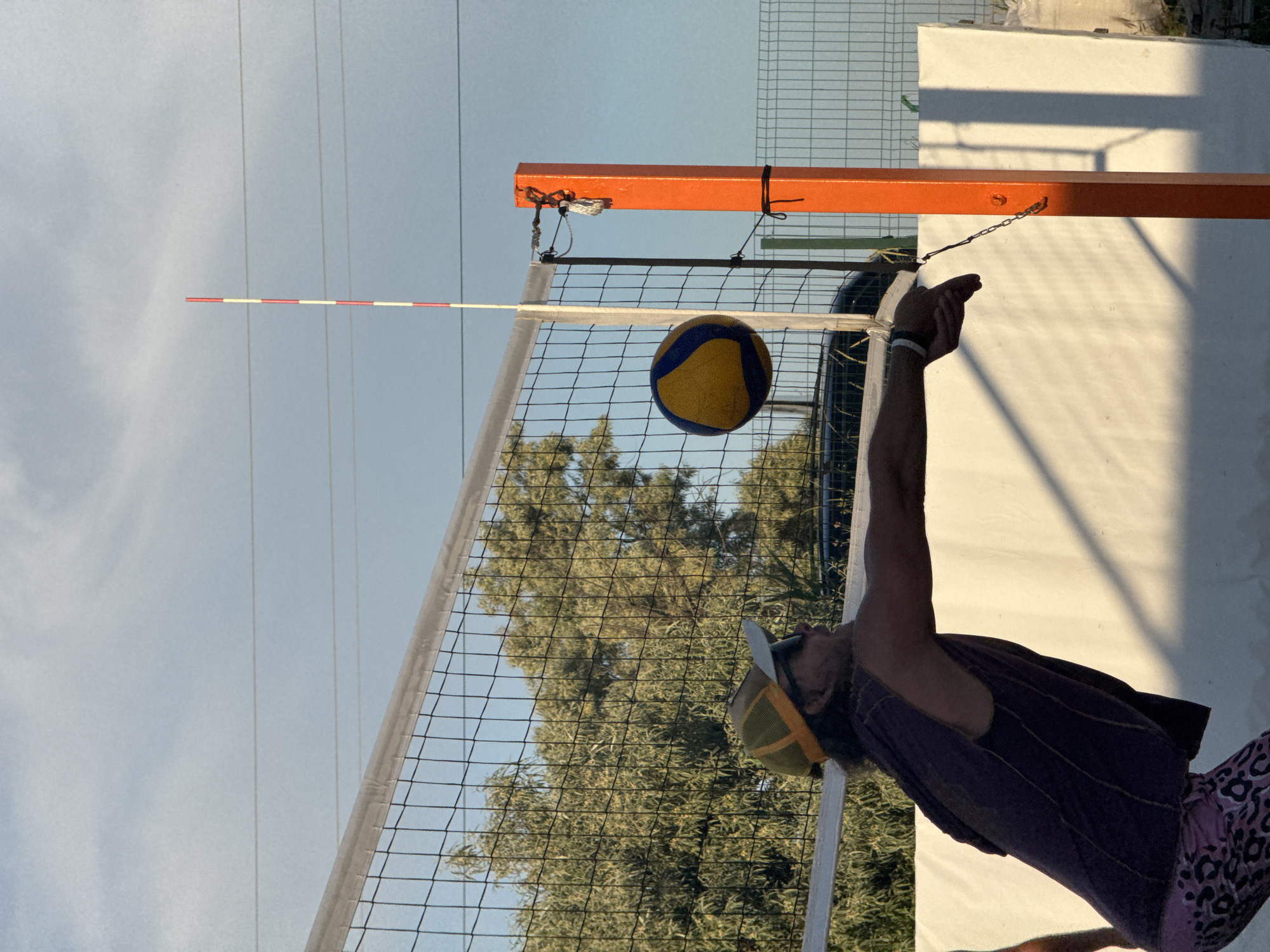 A person is playing volleyball on a sandy court. The person, wearing a yellow and green cap, sunglasses, and a sleeveless purple shirt with a hood, is in the process of serving the ball. The ball, a yellow and blue volleyball, is in mid-air, just above the net. The net is stretched taut and secured to an orange post. Behind the net, trees are visible, and in the background, there is a white wall and a fence. The sky is partly cloudy. The image appears to be taken during daylight. The scene suggests an outdoor recreational activity.