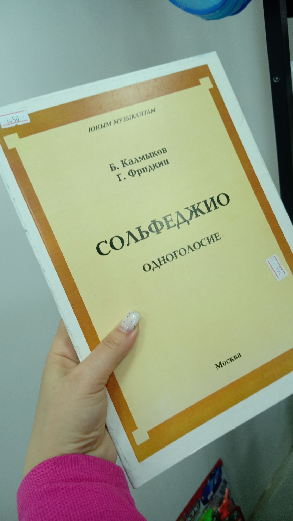 A hand holds a book with a yellow cover and brown border. The text on the cover reads:
ЮНЫМ МУЗЫКАНТАМ (FOR YOUNG MUSICIANS)
Б. Калмыков
Г. Фридкин
СОЛЬФЕДЖИО (Solfege)
ОДНОГОЛОСИЕ (monophony)
Москва (Moscow)
The hand is wearing a pink sleeve and has white nail polish with small black dots. A price tag of 1450 is visible on the book's upper left corner. The book is being held against a white wall. Part of a shelf is visible in the background. The location in the city of Unknown, Unknown is unspecified.