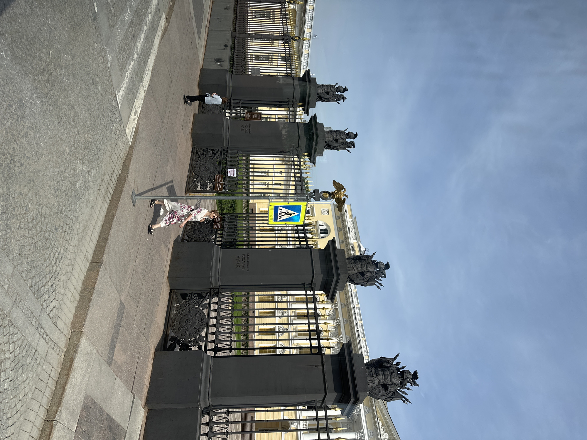 The image shows a street scene in Saint Petersburg, Russia. A row of black, ornate iron gates with decorative elements line the sidewalk. These gates likely enclose a significant building or area. The background reveals a multi-story building with a yellow facade and numerous windows. A pedestrian wearing a white dress with floral patterns walks along the sidewalk beside the gate. Another person in a white shirt and black pants is standing further down the sidewalk near another segment of the gate. A blue and yellow pedestrian crossing sign is visible near the foreground. The sky is a clear blue. The image is taken during daylight hours, with the sun casting shadows on the scene. The ground is composed of paved tiles.