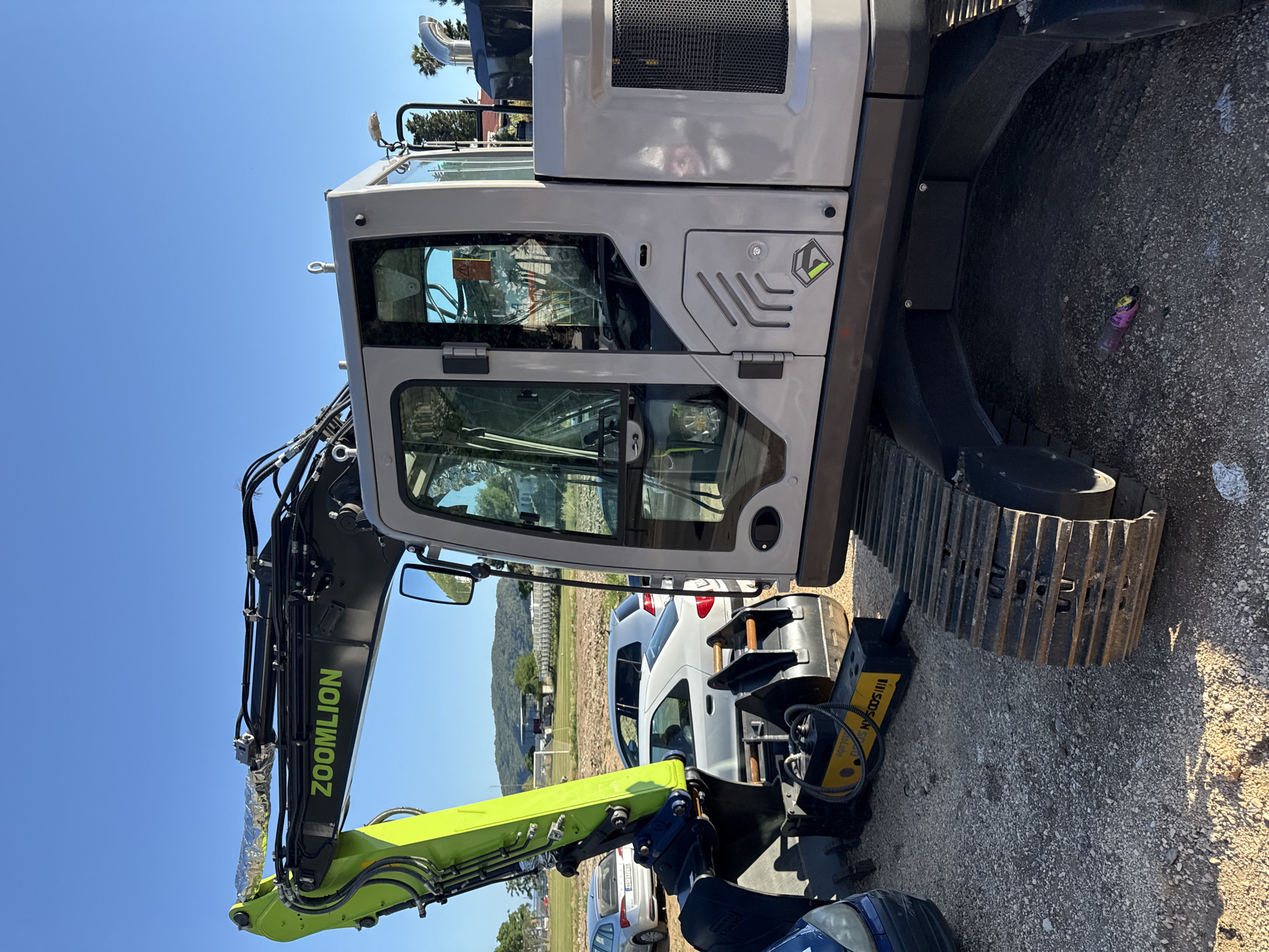 The image displays a Zoomlion tracked excavator parked on a gravel and dirt surface under a clear blue sky in Budva, Montenegro.
The excavator features a prominent lime green boom with "ZOOMLION" branding in black lettering. Its main body and cab are light grey, with a black-grilled engine compartment visible on the right side. Hydraulic hoses run along the boom, one segment wrapped in metallic foil. A yellow and black Soosan hydraulic breaker attachment is affixed to the excavator's arm. The tracks are dark grey and show signs of wear. Underneath the excavator, a discarded pink plastic bottle is visible.
In the mid-ground, a person wearing dark clothing is partially visible, bent over near the front of a blue car on the far left. Several other vehicles are parked nearby: a silver Skoda Octavia with a Serbian license plate "BG-2923 EC", a white sedan, and a silver Volkswagen Golf.
The background reveals a football field with a white goalpost and painted lines. Behind the field, various buildings and structures are discernible. Further in the distance, green hills and mountains rise under the clear sky. Palm trees are visible on the right side of the frame, suggesting a warm climate. The scene is consistent with an open area, possibly near a stadium or sports complex, within the coastal city of Budva.