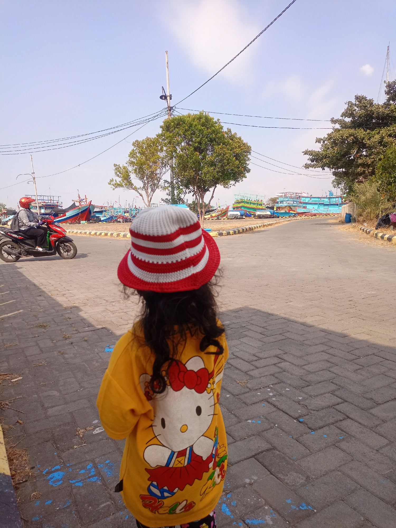 In Juwana, Indonesia, a young child wearing a red and white striped bucket hat and a yellow Hello Kitty shirt stands on a paved walkway. The child's back is to the camera, and they are looking out towards a street where a person on a red motorcycle is riding past. In the background, numerous fishing boats are docked in what appears to be a harbor or boatyard. Trees line the side of the street, and utility poles with wires are visible against a clear blue sky.