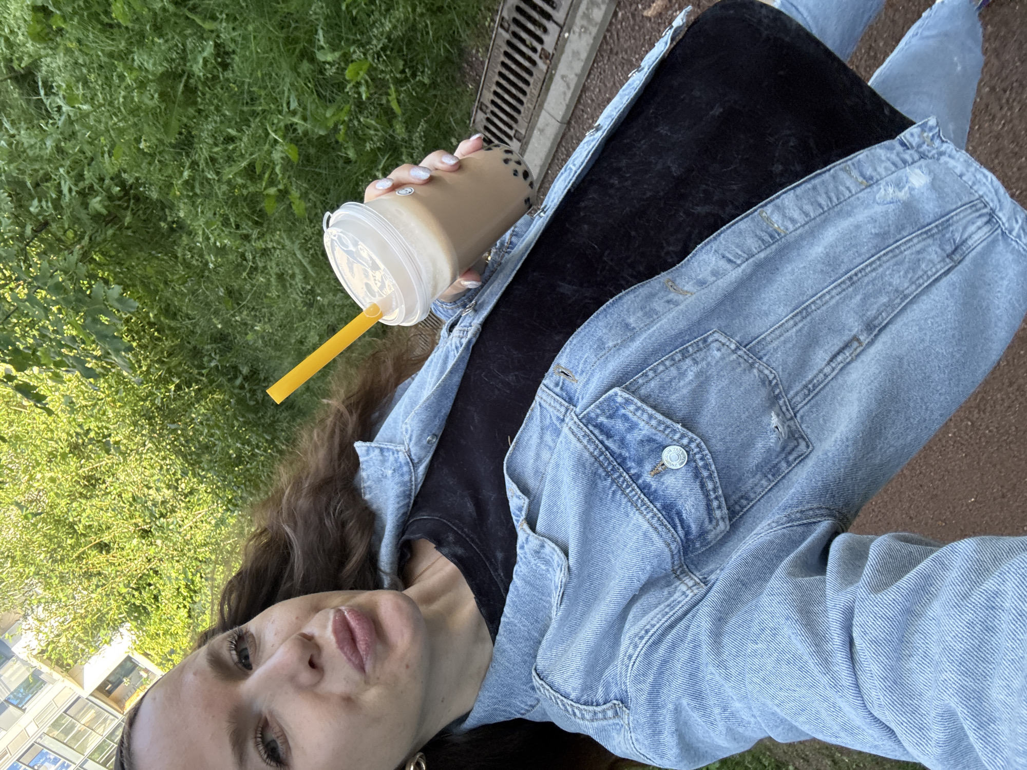 In this media file, a young woman is captured from the chest up, holding a bubble tea. She is wearing a light blue denim jacket over a black t-shirt and is outdoors with green grass and a building visible in the background. A drainage grate is also present near her. The woman has long, dark, wavy hair and is looking towards the camera with a slight smile. Her nails are painted with a glittery polish. The time of day appears to be daytime, with bright lighting. The context provided places the scene in Moscow, Russia.
