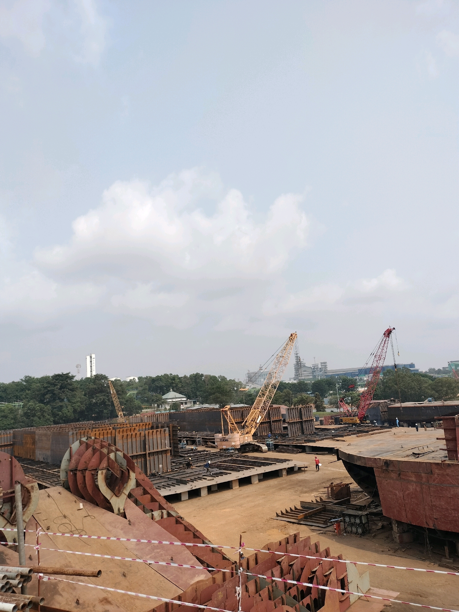 This image depicts a shipyard in Batam, Indonesia. The scene is dominated by the construction of large vessels, with numerous metal components and partially assembled ship sections visible. Two prominent cranes, one yellow and one red, are positioned within the yard, suggesting ongoing heavy lifting operations. Several workers, dressed in casual attire, are scattered throughout the site, some seemingly engaged in work while others are walking. In the background, a line of trees separates the shipyard from industrial buildings and a tall, white tower-like structure. The sky is overcast with scattered clouds, and the ground is a light brown, sandy material. The foreground features stacked metal pipes and warning tape.