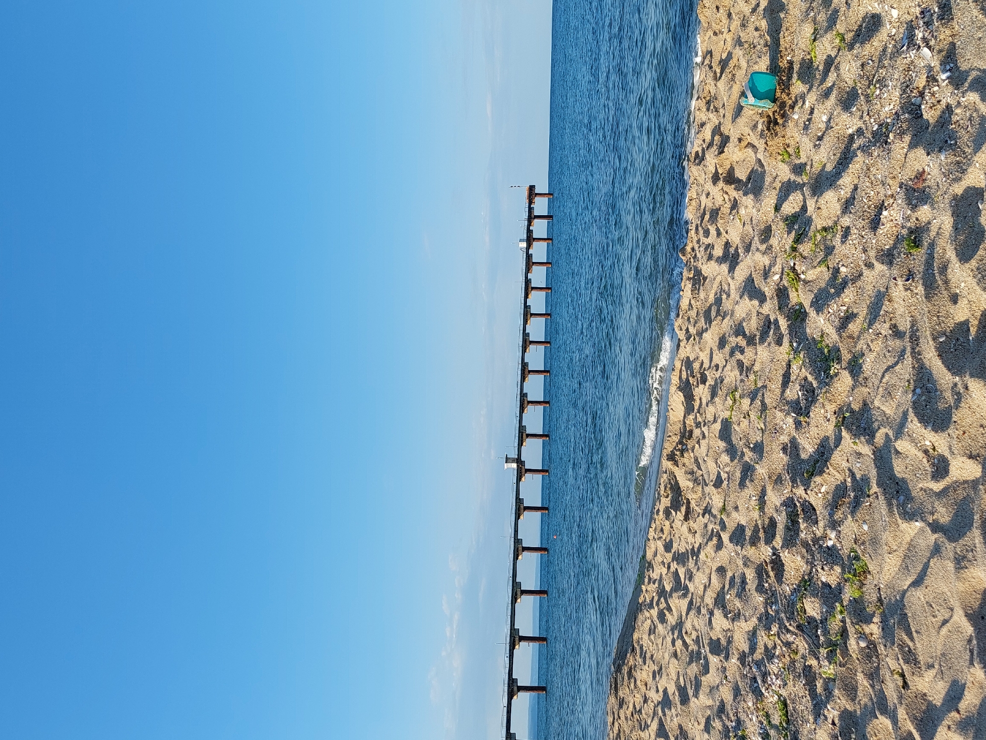A long pier extends from the left side of the image, stretching across the blue sea towards the right horizon. The pier is supported by multiple pilings and features a small, white building structure in the middle. The water beneath the pier is calm with small waves lapping at the shore. The foreground is a sandy beach with shells and scattered green vegetation. On the right side of the beach, a small, teal-colored plastic bucket lies on its side. The sky is a clear, bright blue with a few wispy white clouds visible near the horizon. This scene depicts the beach and pier in Shkorpilovtsi, Bulgaria.
