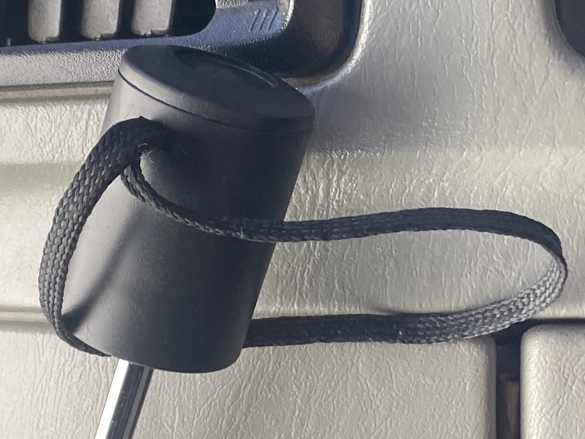 In Rozhny, Ukraine, an object, possibly a black cylindrical knob with a braided cord attached, is fastened to a metallic rod protruding from a light-colored, textured surface. The knob is dark and matte, with the cord wrapped around its circumference and hanging loosely downwards. To the right of the knob is a section of dark material with horizontal, parallel grooves, suggesting it might be part of a vehicle's interior or a similar manufactured item. The overall scene is brightly lit, casting subtle shadows and highlighting the textures of the surrounding surfaces.
