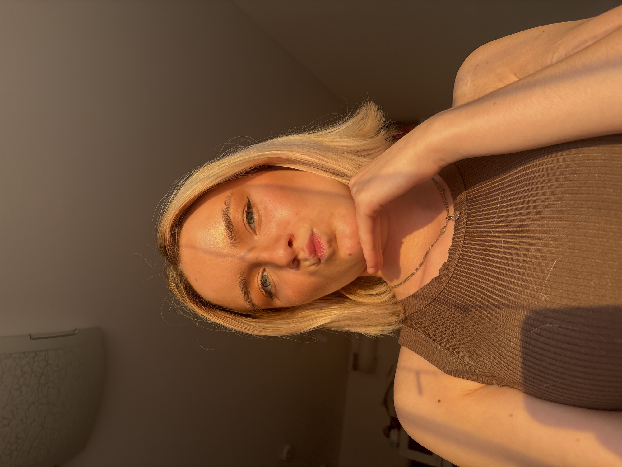The image features a young woman posing for a close-up, rotated 90 degrees clockwise. She has blonde, chin-length hair and fair skin, illuminated by warm, golden light that suggests late afternoon or "golden hour." Her eyes are light-colored, possibly green or blue, and she wears subtle eyeliner. Her lips are slightly pursed in a pout.
She is wearing a ribbed, brown sleeveless top and a thin silver necklace with a small, circular pendant. Her left hand is resting on her chin or jawline. The lighting creates distinct highlights on her skin and hair, with soft shadows.
The background consists of plain, neutral-colored walls, darker on the right side and lighter on the left. A light fixture or decorative element is partially visible and out of focus on the far left. The overall impression is an informal, indoor portrait, likely a selfie. No text is visible in the image.
