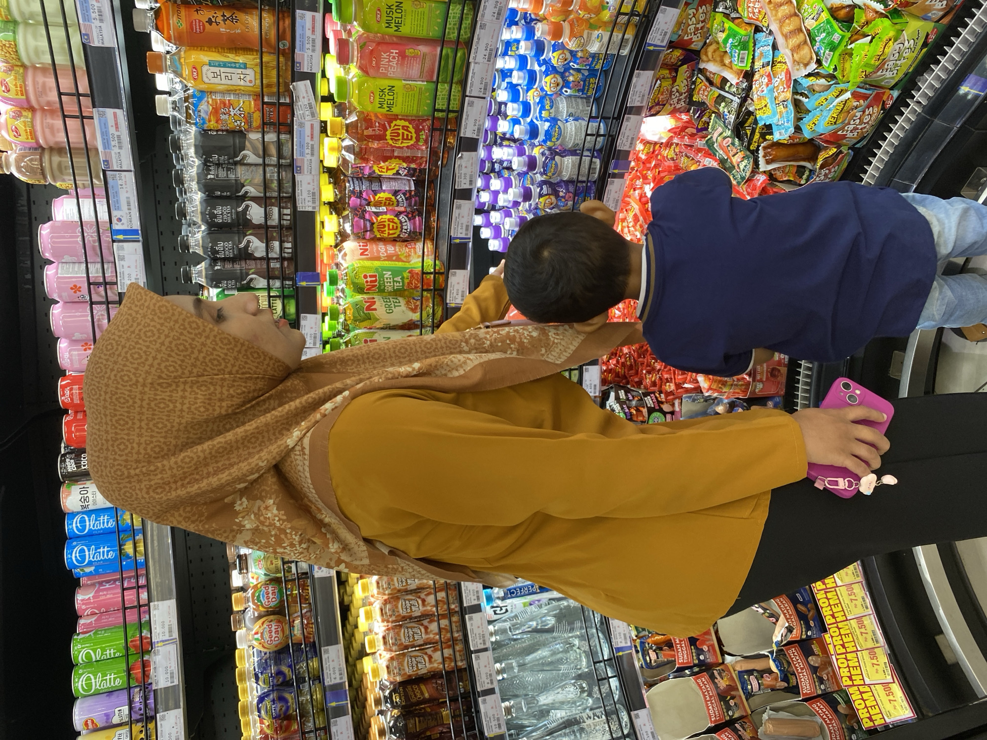 In a brightly lit supermarket aisle in Banda Aceh, Indonesia, a woman wearing a mustard yellow long-sleeved shirt and a patterned brown hijab is browsing drinks on a shelf with a young boy, likely her son, who is wearing a dark blue t-shirt and light blue jeans. The woman is standing and looking to her right at the beverage selection, while the boy is facing away from the camera, looking towards the shelves with a focused gaze.
The shelves are filled with a variety of beverages, including cans of "latte" in various colors, bottles of "Good Day" coffee, and bottles of tea like "OKF Green Tea" and "OKF Fruit Tea." To the right of the woman, there are rows of small bottles with purple and blue liquid, some labeled "Pororo." Further down the aisle, the boy is positioned in front of shelves stocked with packaged snacks, predominantly brightly colored bags of chips and candies. One noticeable item in the boy's direct line of sight is a package of sausages.
The woman is holding a bright pink smartphone with a cloud-shaped keychain in her left hand. The aisle appears to be well-stocked, with clear pricing labels visible on some of the products, indicating typical supermarket displays. The overall environment suggests a normal shopping trip for groceries and beverages.