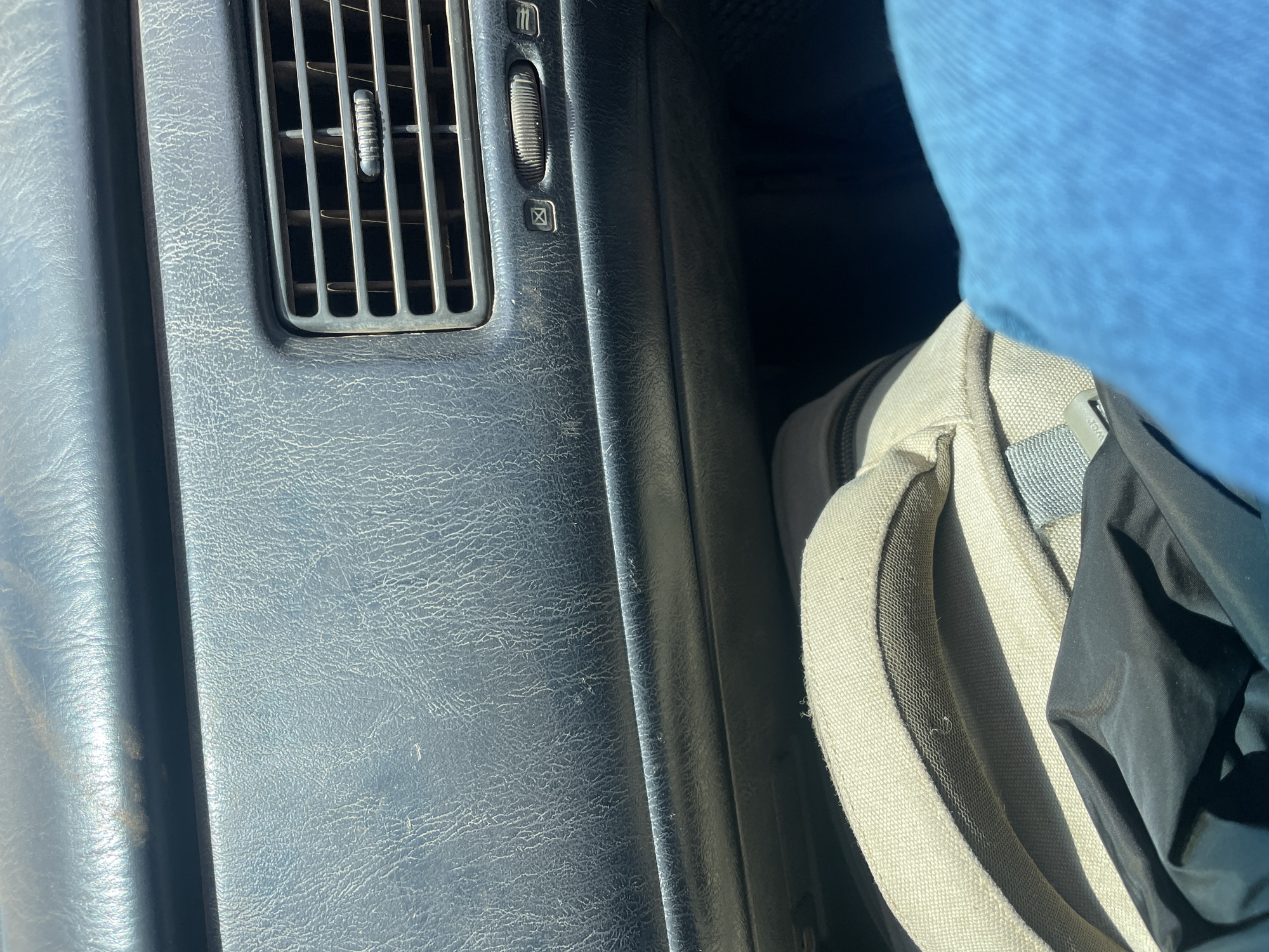 The image shows the dashboard of a vehicle. The dashboard is textured and has an air vent with adjustable louvers and a dial. Next to the dashboard is a backpack with a tan strap and some black material. A blue object is visible above the backpack, possibly clothing. The location is unknown and there is no information about Unguwar Gaya, Nigeria, or any people or interactions.